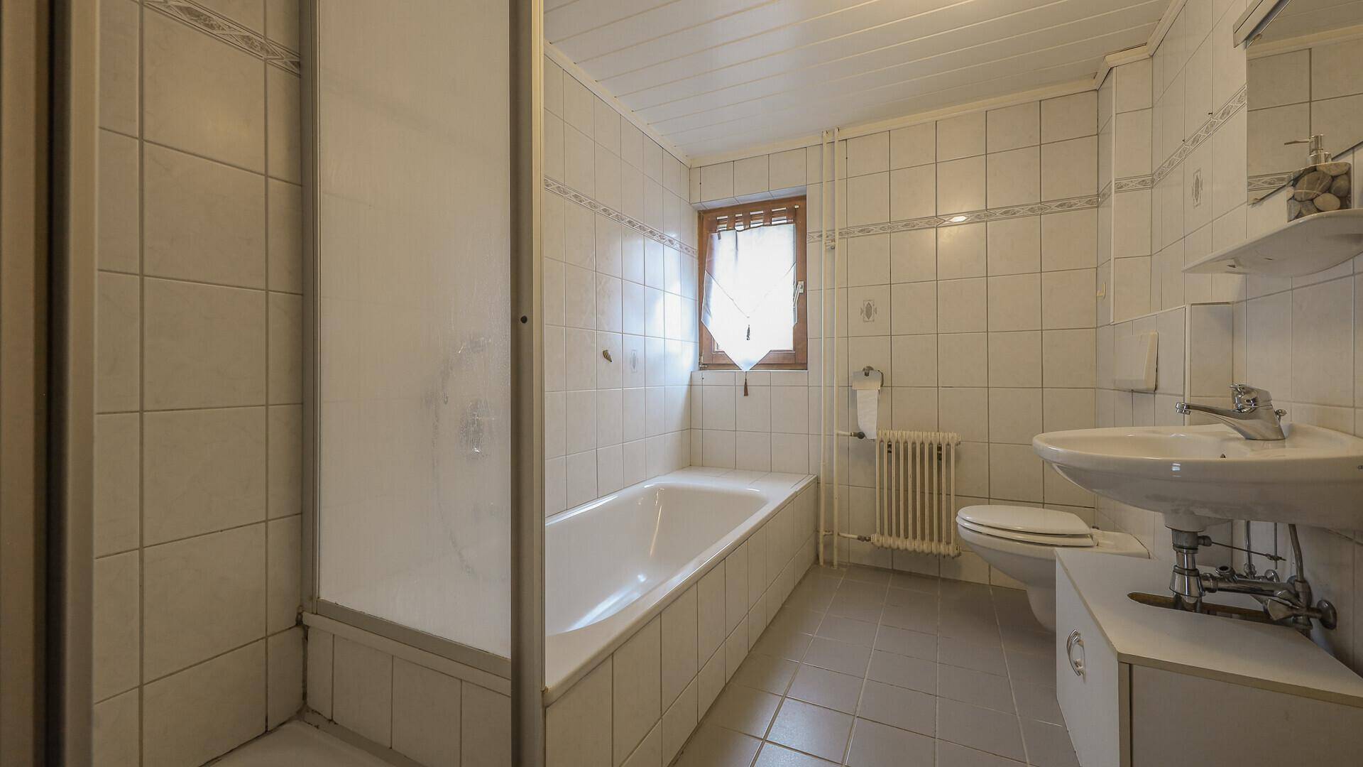 Helles Badezimmer mit Badewanne, Dusche und Fenster, ausgestattet mit weißen Fliesen.