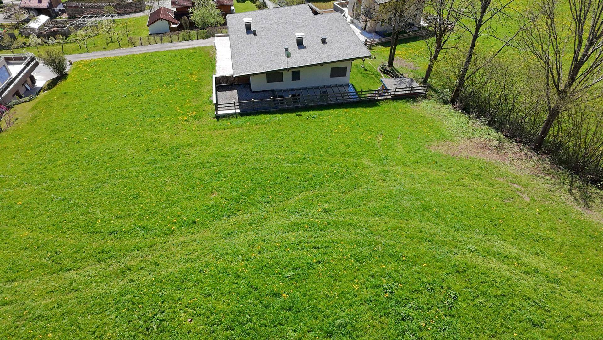 Luftaufnahme eines Hauses mit grauen Dachziegeln und einer Terrasse, umgeben von einer großen Wiese.