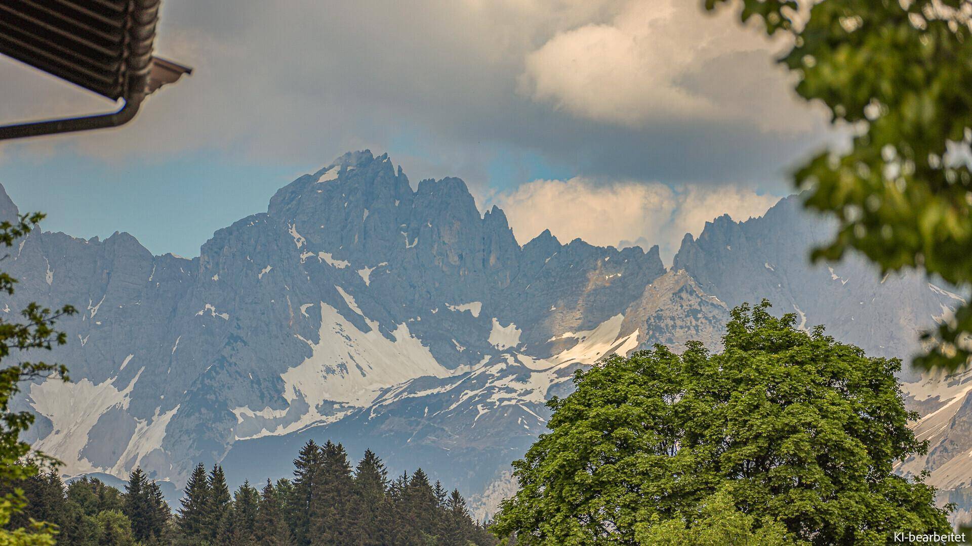 Panoramablick auf schneebedeckte Berge und grüne Bäume unter einem bewölkten Himmel.