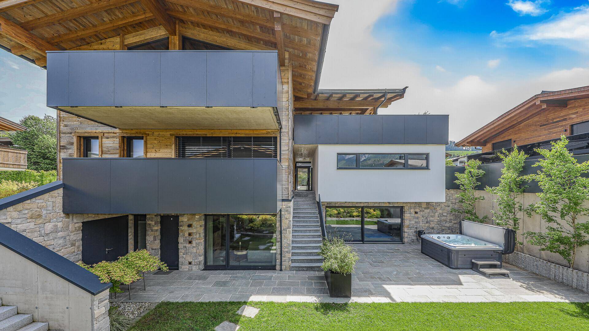 Moderne Villa mit Holz- und Steinfassade, Balkonen und Außentreppe. Ein Whirlpool befindet sich auf der Terrasse.
