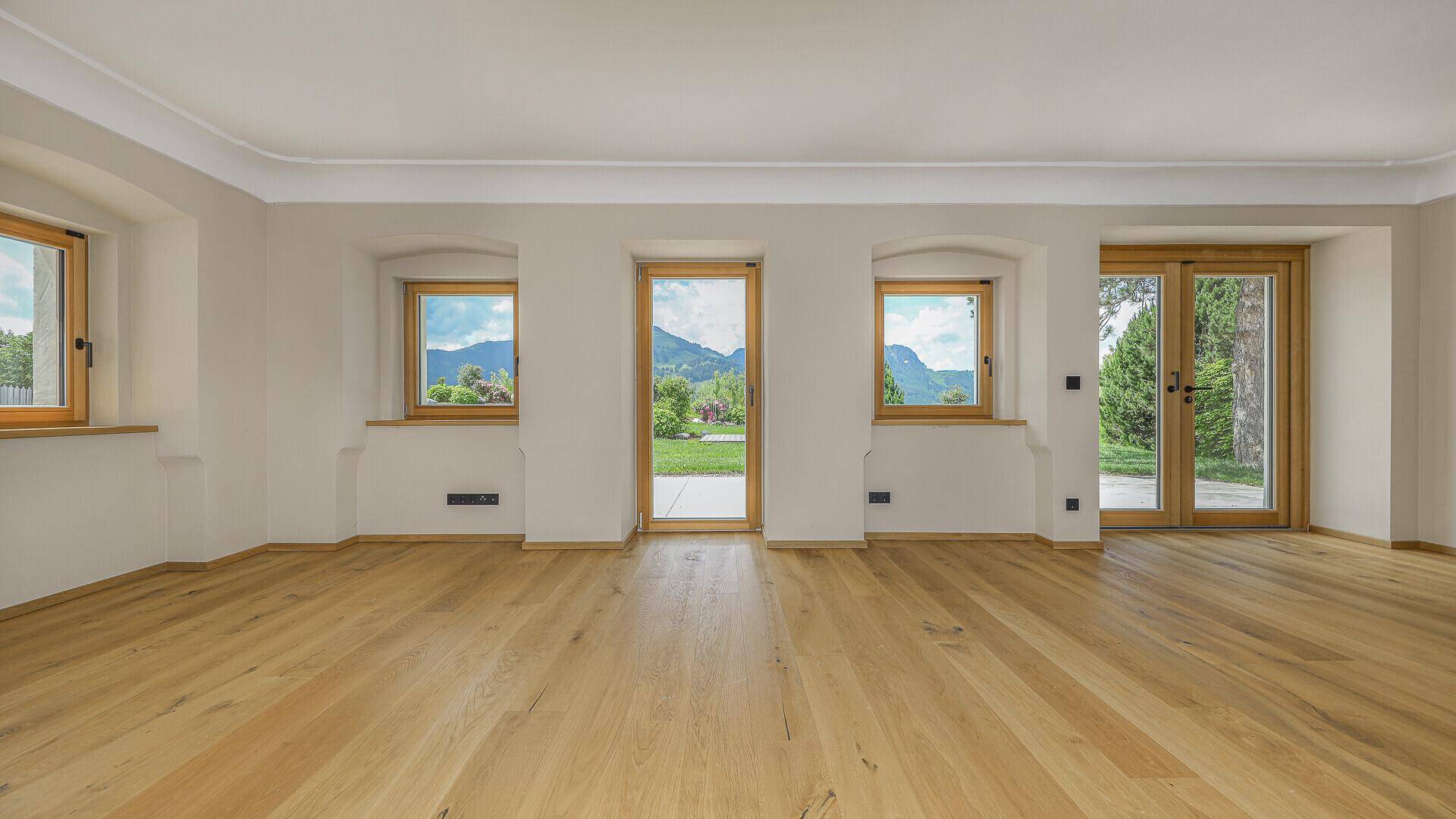 Geräumiger Raum mit Parkettboden, Stuckdecke und mehreren Fenstern mit Gartenblick.