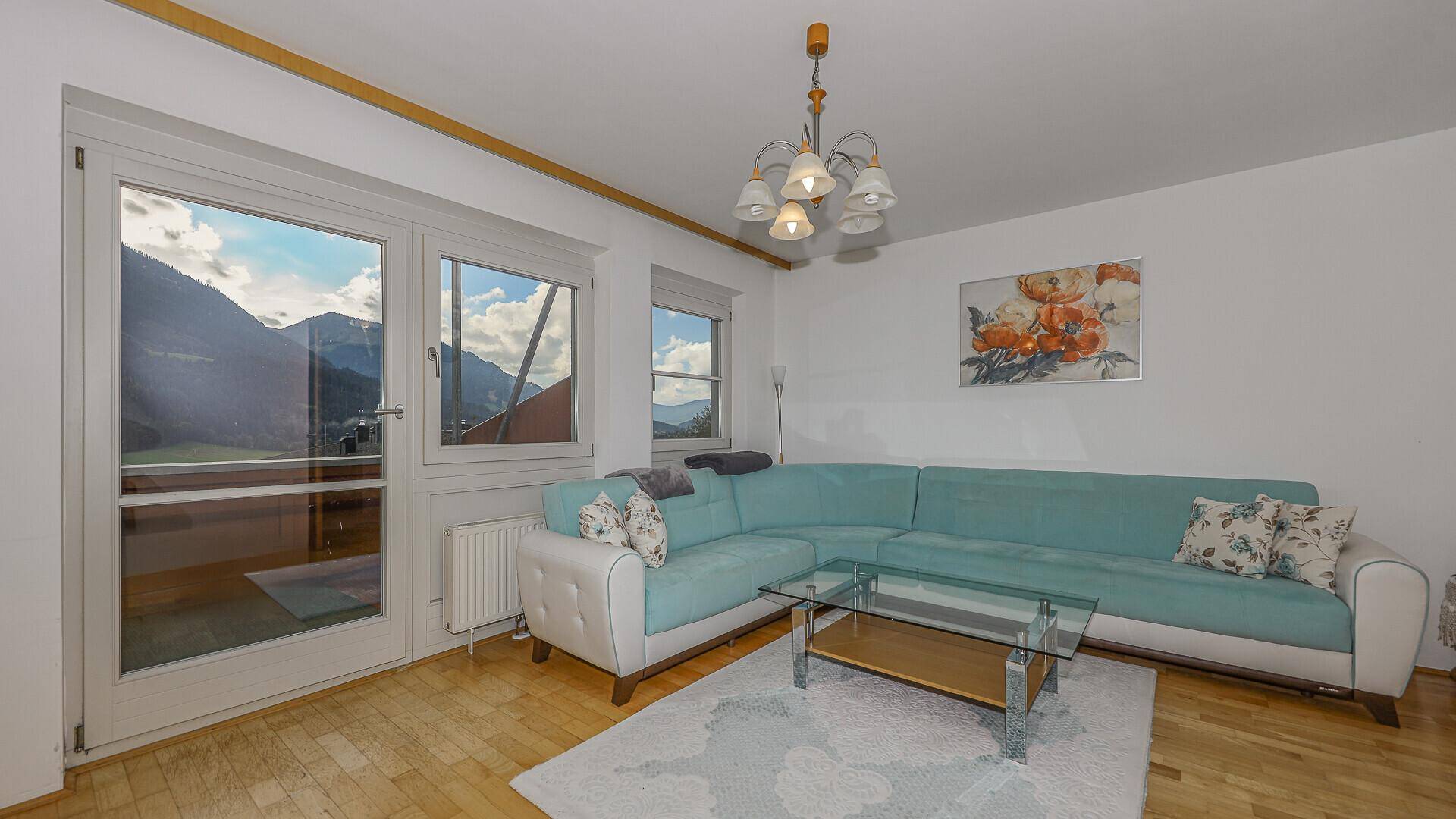 Wohnzimmer mit türkisem Ecksofa, Holzboden und Zugang zu einem Balkon mit Bergblick.
