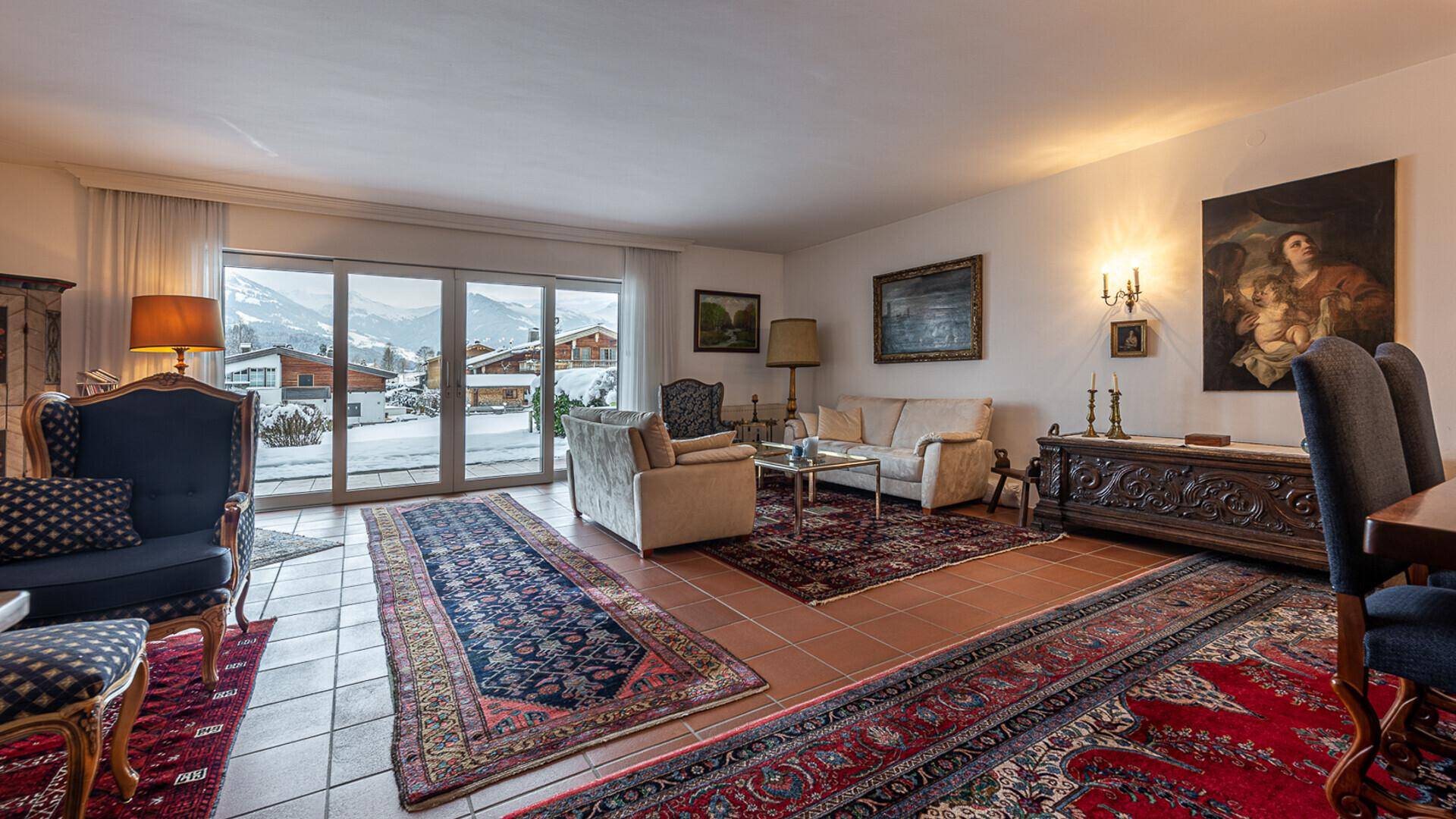 Gemütliches Wohnzimmer mit traditionellen Möbeln, Teppichen und Blick auf die verschneite Berglandschaft.