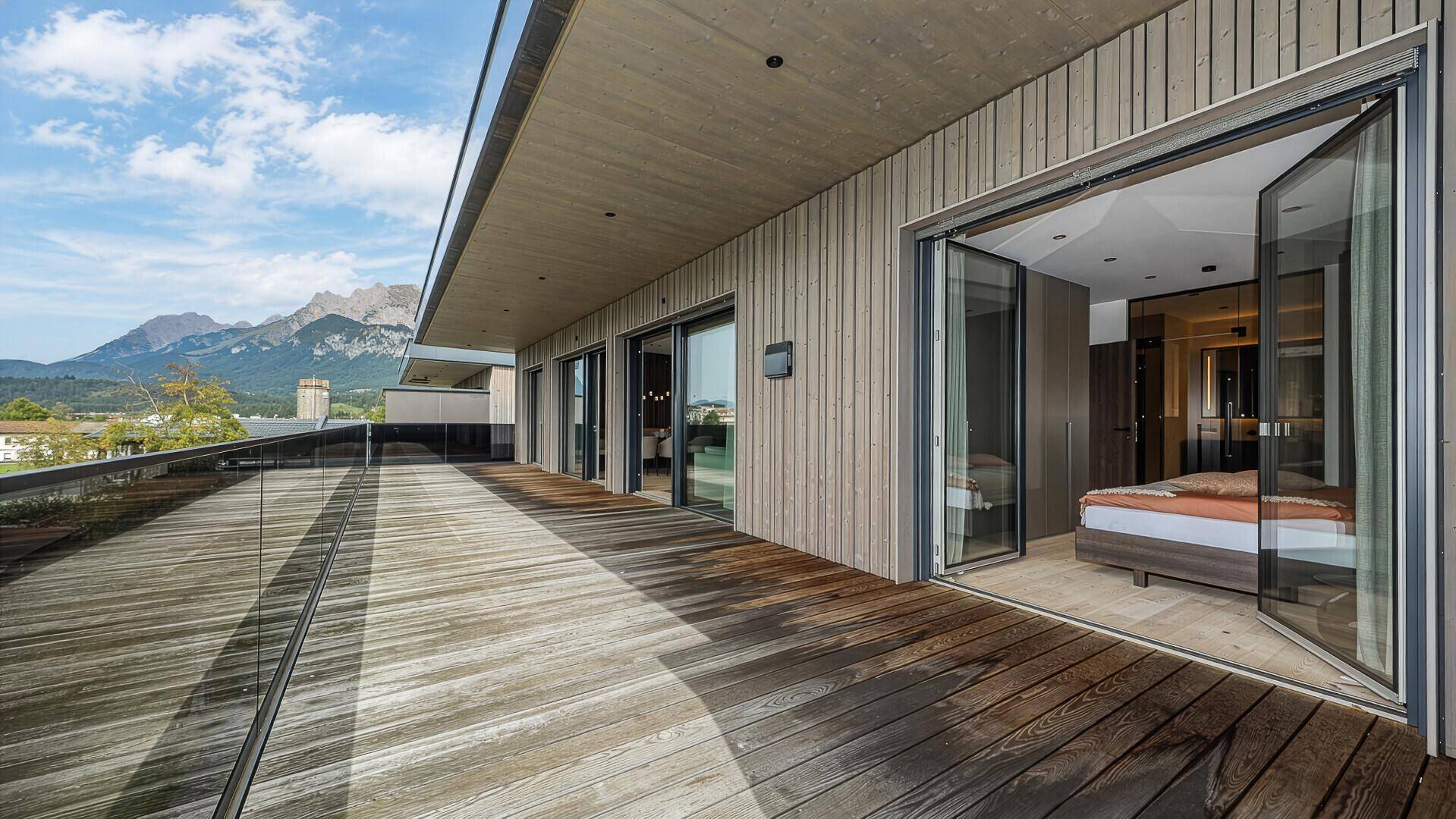 Weitläufige Holzterrasse mit Glasgeländer und Zugang zu einem Schlafzimmer mit Bergblick.