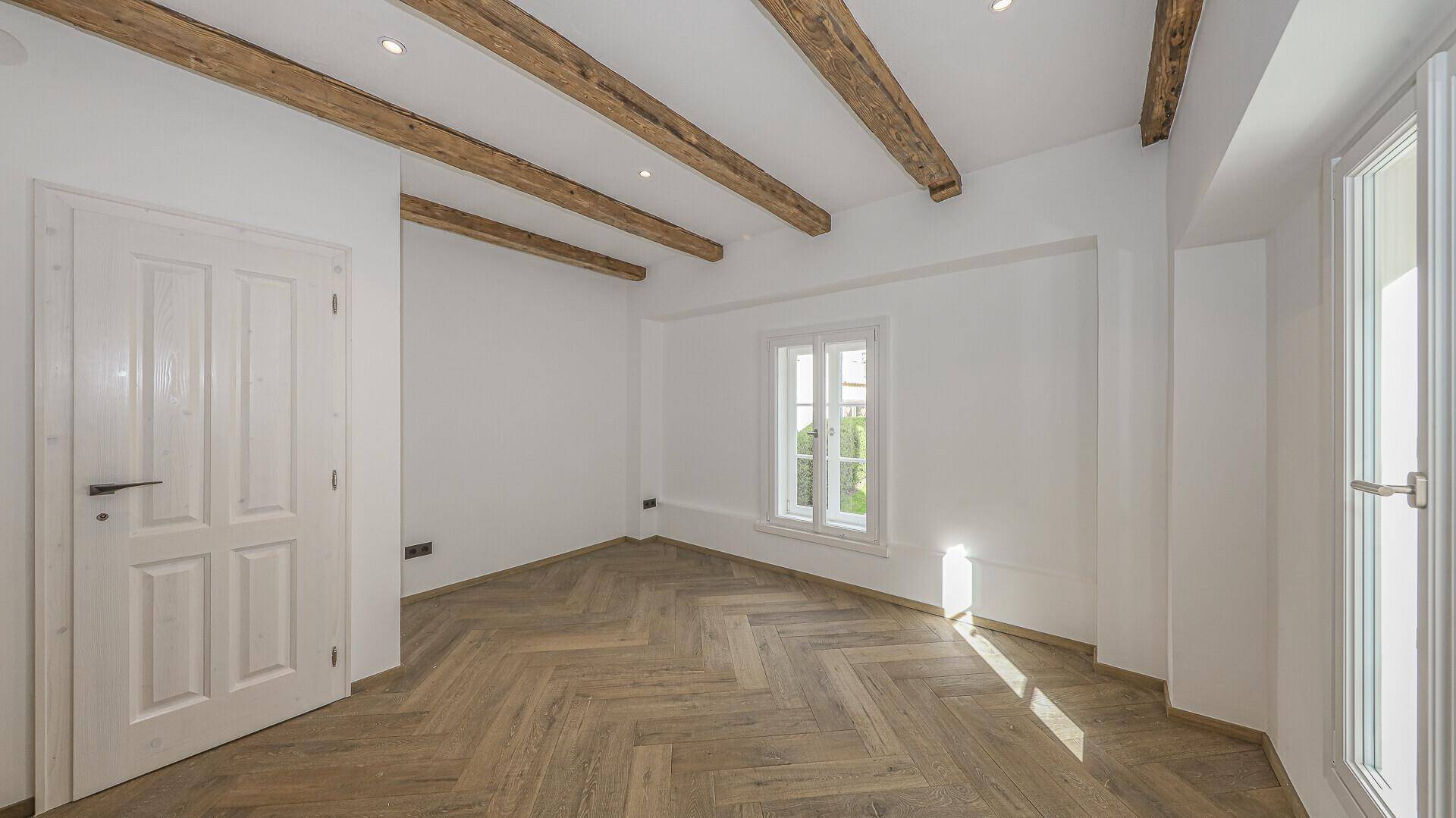 Helles Zimmer mit Fischgrätparkett, weißen Wänden und sichtbaren Holzbalken.