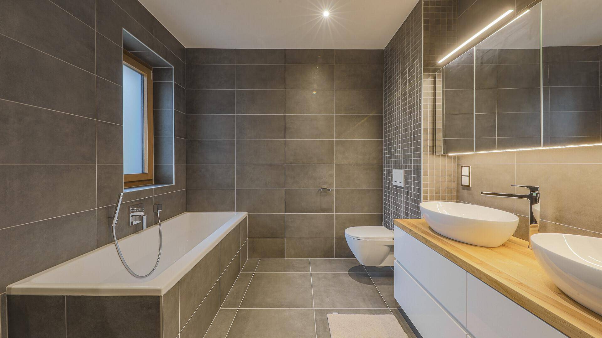Modernes Badezimmer mit grauen Fliesen, Badewanne, Doppelwaschbecken und Fenster.