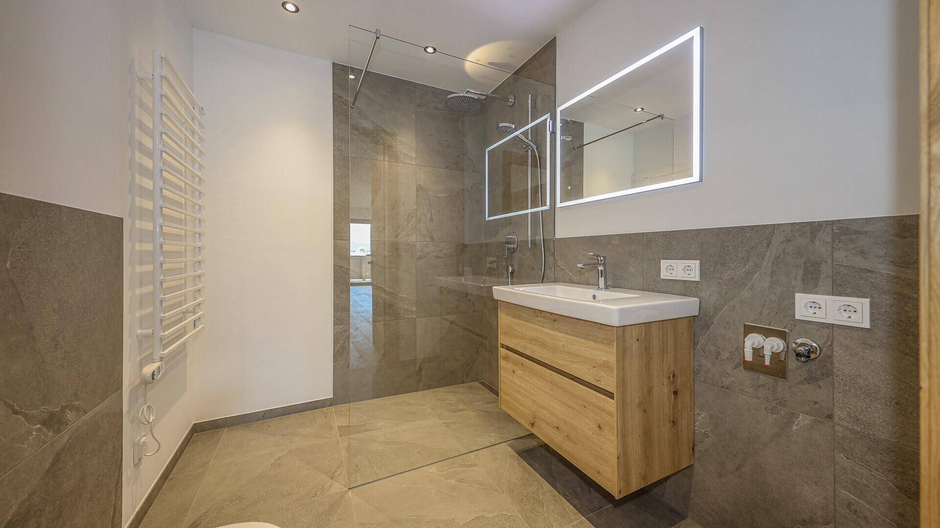 Modernes Badezimmer mit begehbarer Dusche, Holzwaschtisch und beleuchtetem Spiegel.