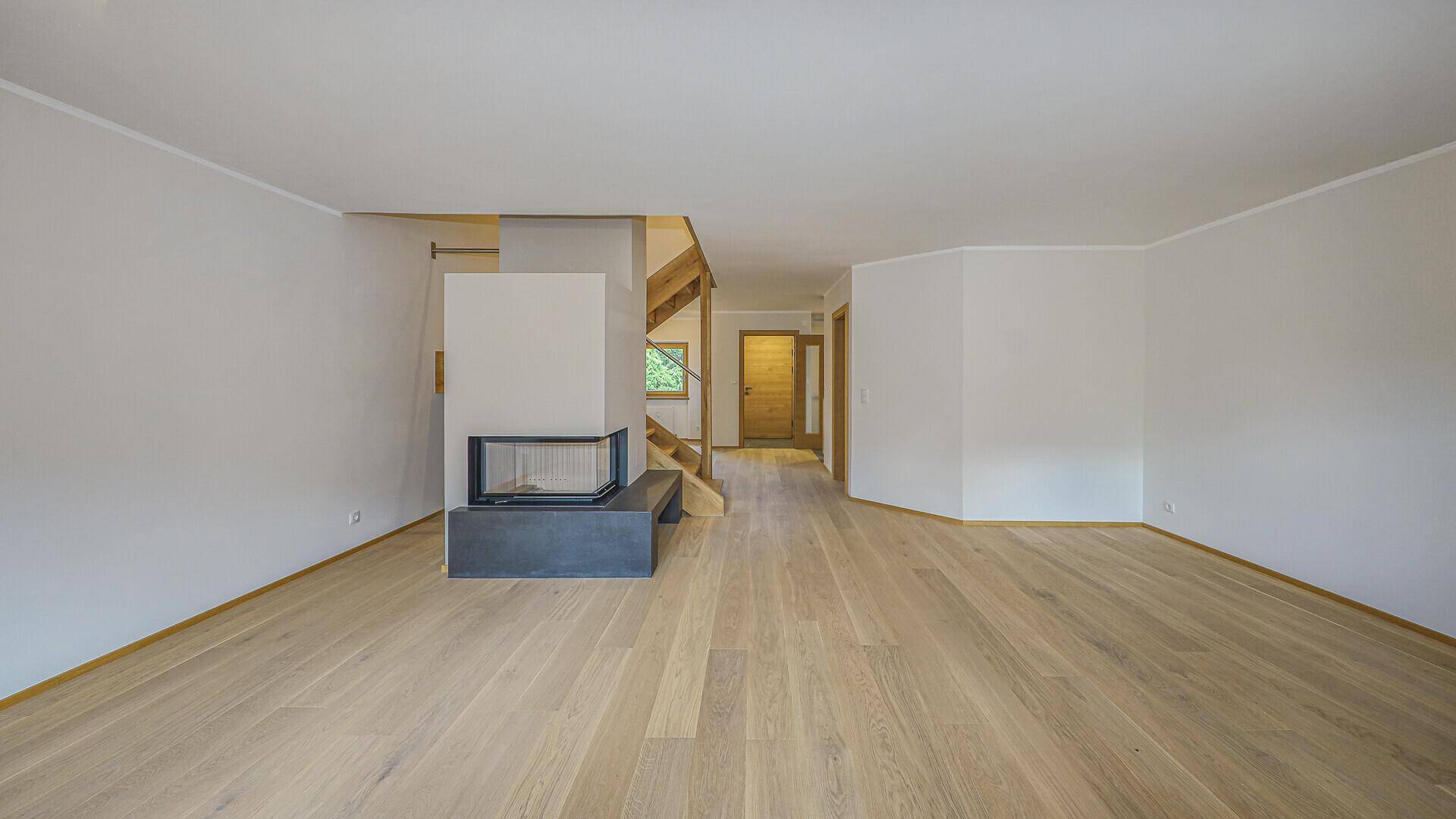 Leeres Wohnzimmer mit hellem Holzboden, modernem Kamin und offener Holztreppe zum Obergeschoss.