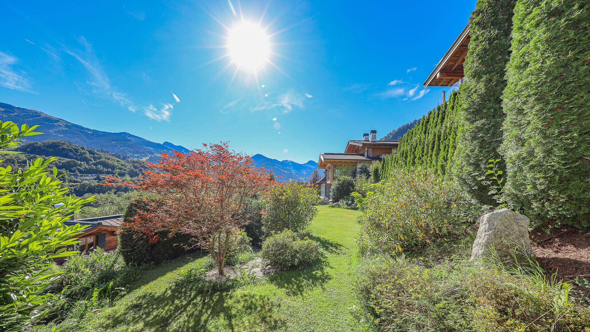 Sonnige Gartenlandschaft mit üppiger Bepflanzung und Blick auf die umliegende Bergwelt und das Haus.