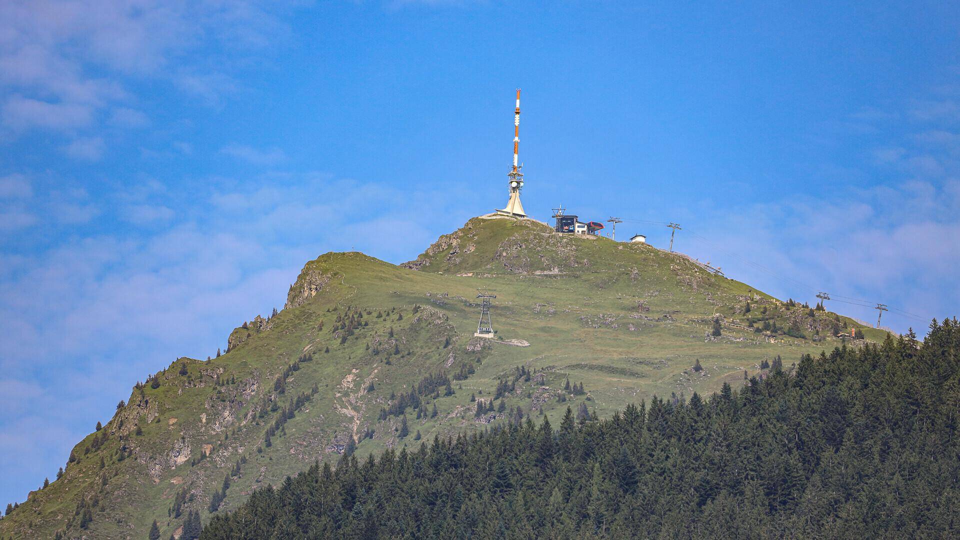 Gipfel eines grünen Berges mit einer Antenne und Seilbahnstation unter klarem blauem Himmel.