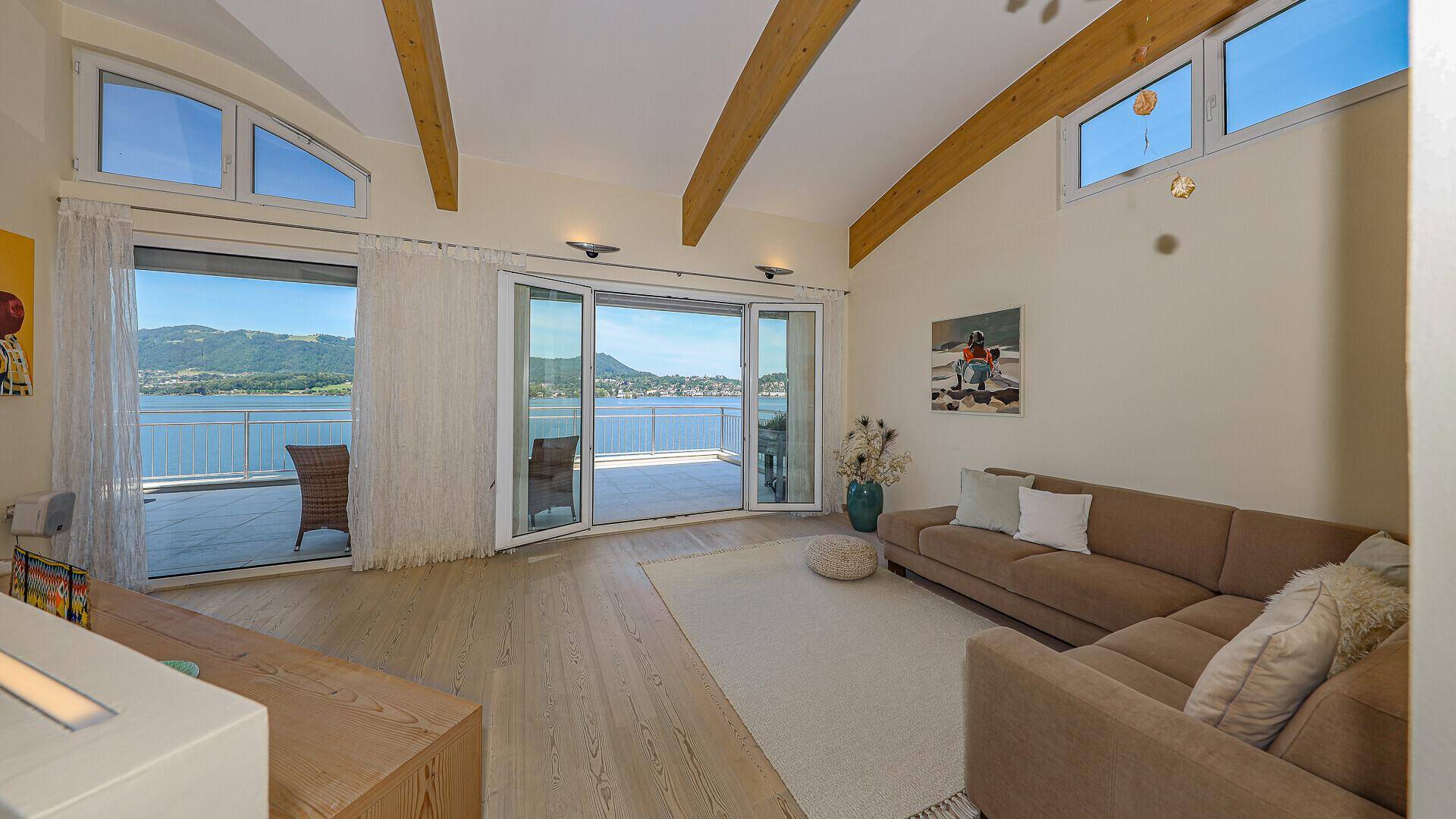 Geräumiges Wohnzimmer mit Holzbalken, großen Fenstern und direktem Zugang zur Seeblick-Terrasse.