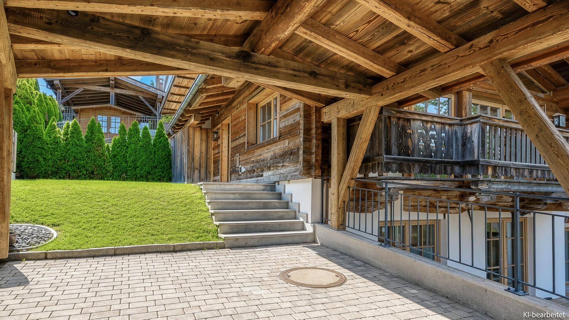 Überdachter Carport mit gepflasterter Einfahrt und Treppe zum Holzhaus, umgeben von Grünflächen.