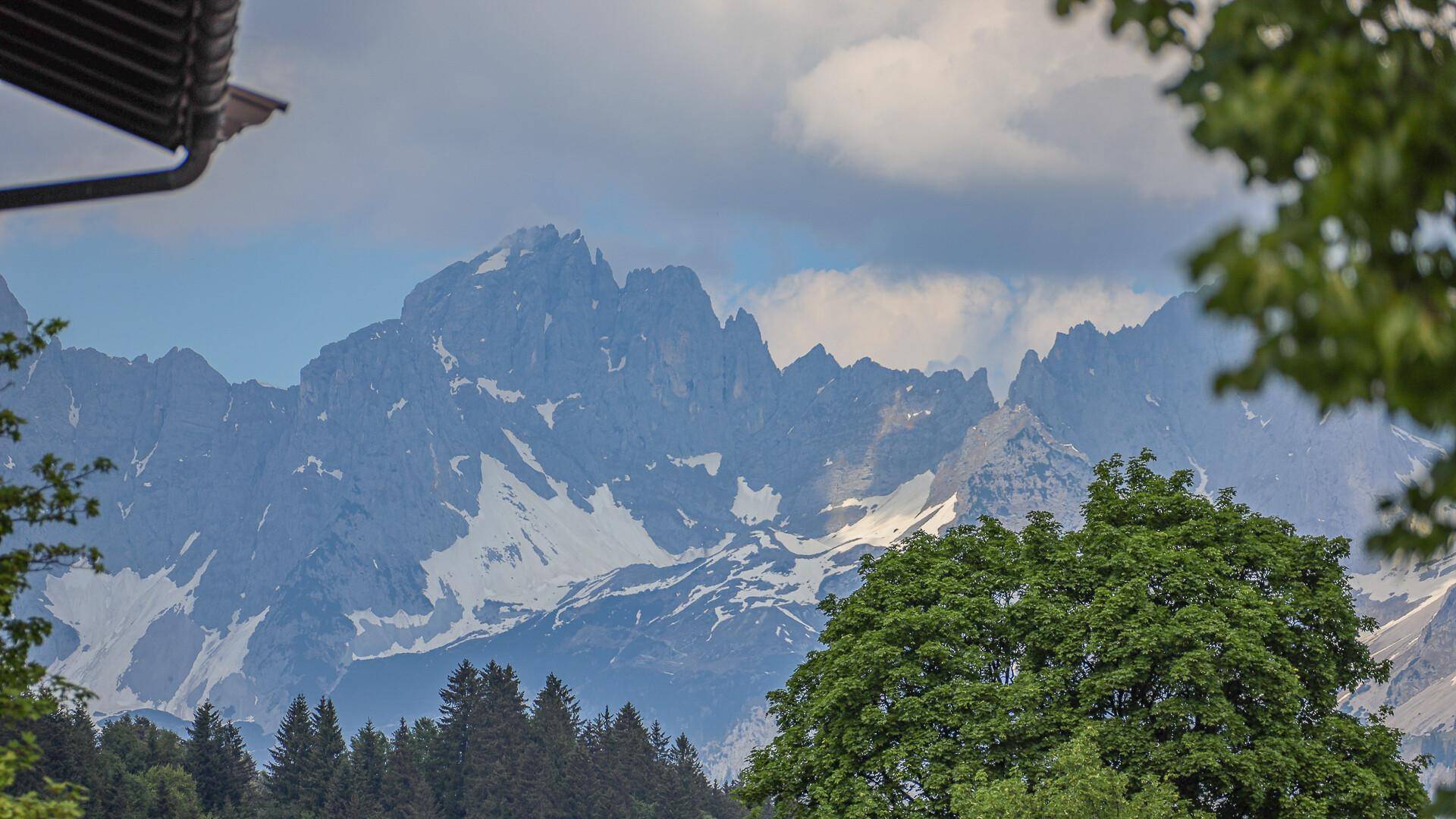 Majestätischer Bergblick mit schneebedeckten Gipfeln und grünen Wäldern unter bewölktem Himmel.