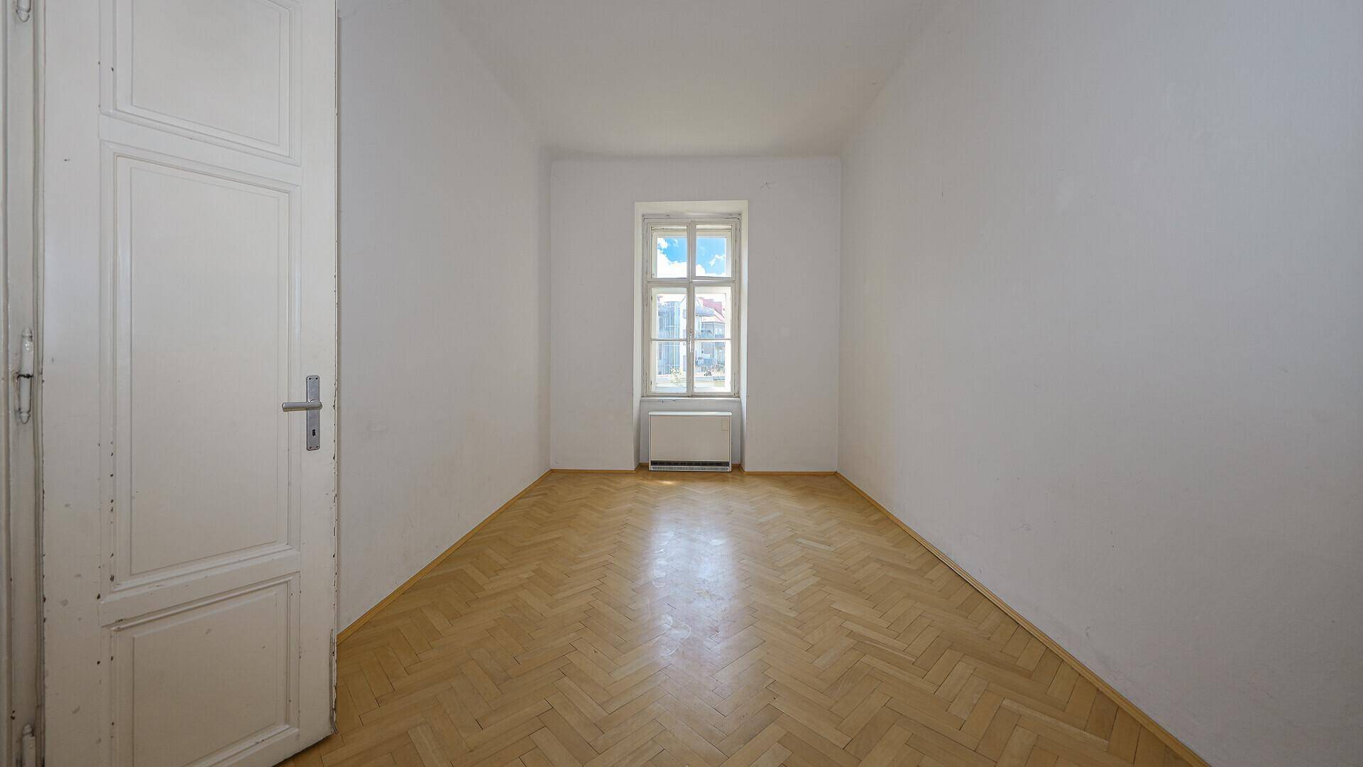 Schmaler Raum mit Parkettboden, einem Fenster und einer hohen Tür.