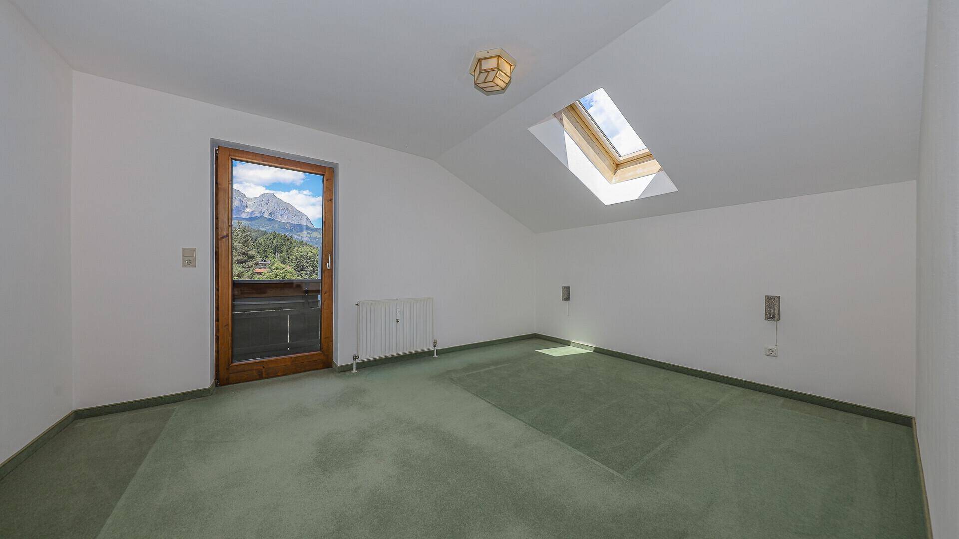 Leeres Schlafzimmer mit grünem Teppichboden, Dachfenster und Balkonzugang mit Bergblick.