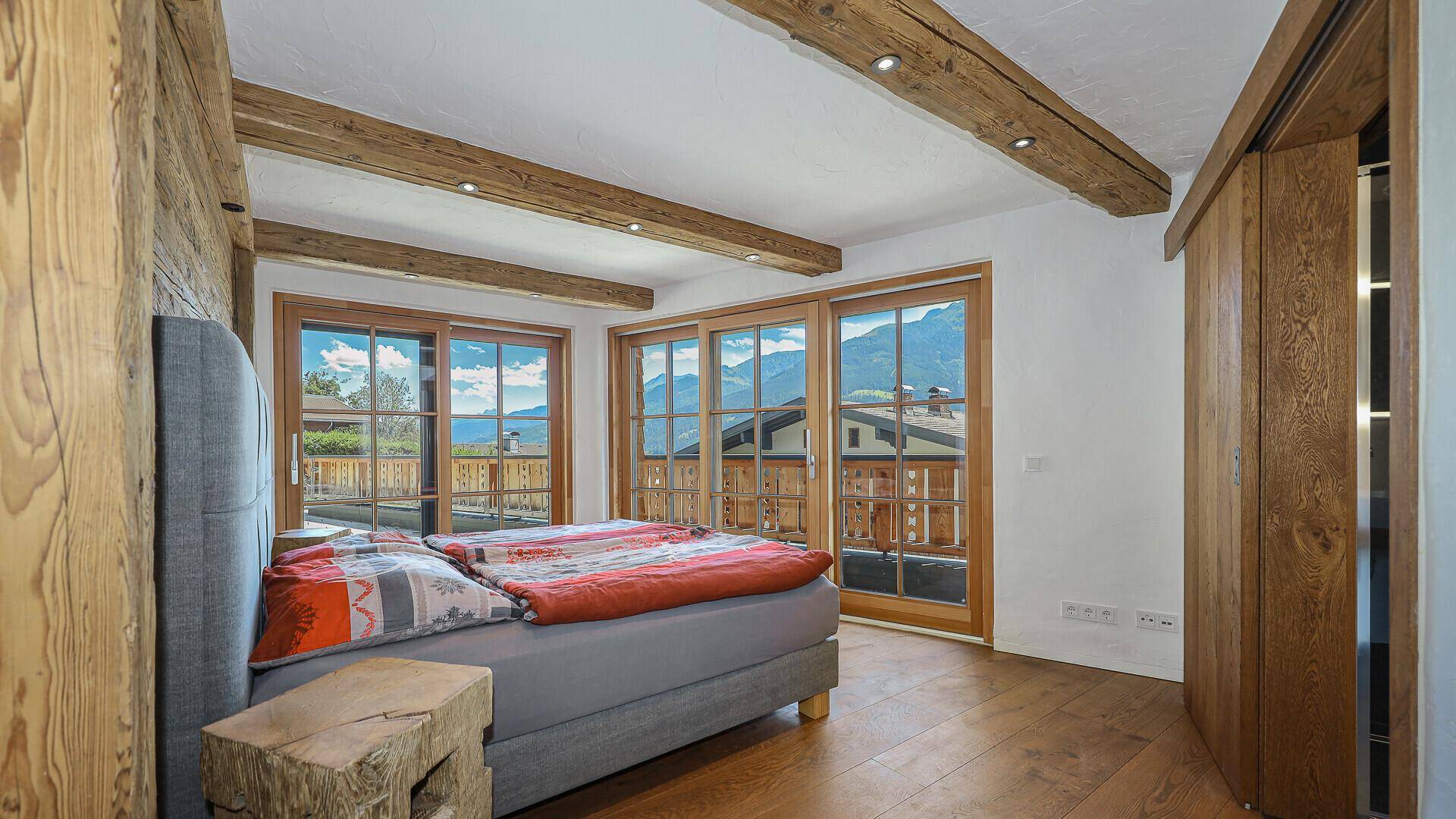 Helles Schlafzimmer mit Holzbalken, großzügigen Fenstern und Zugang zum Balkon.