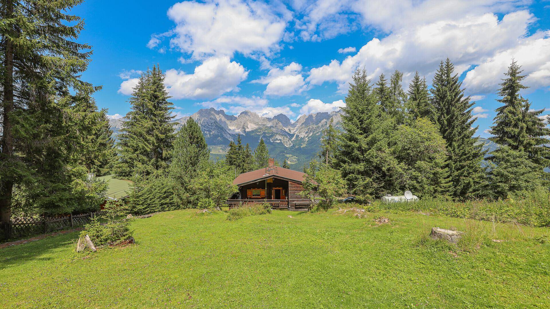 Traditionelles Holzhaus auf einer grünen Wiese mit majestätischem Bergblick unter blauem Himmel.
