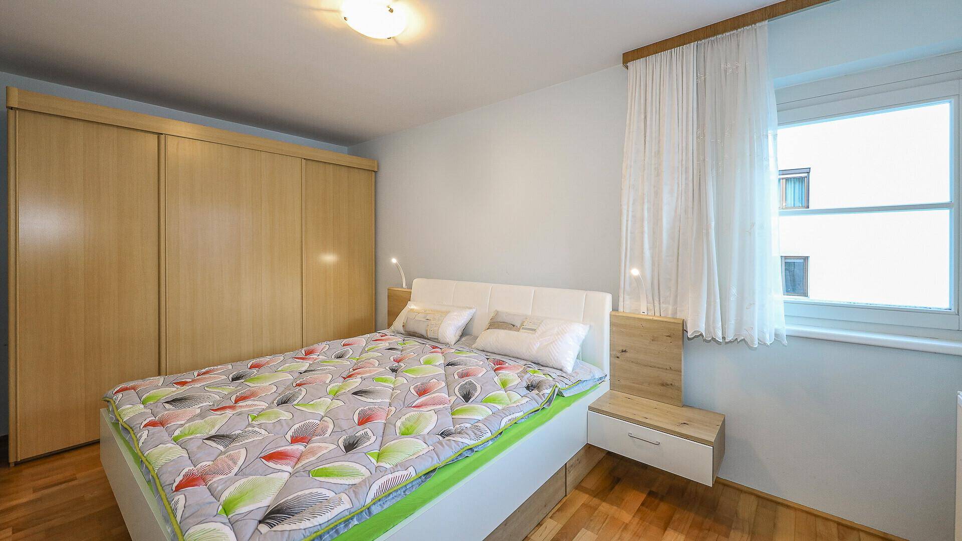 Schlafzimmer mit Doppelbett, Holzschrank und Fenster, einfach und funktional eingerichtet.