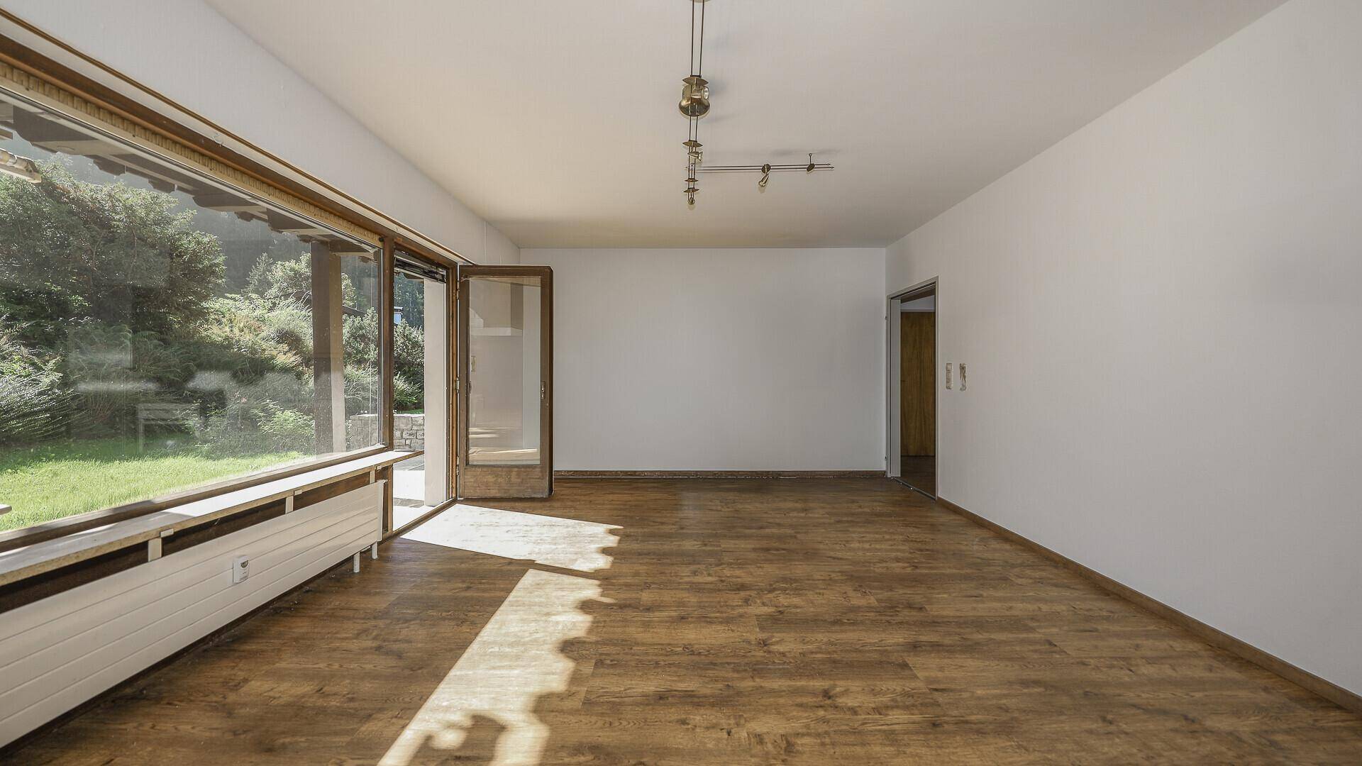 Heller, leerer Raum mit Holzboden und direktem Zugang zum Garten durch große Fenster.
