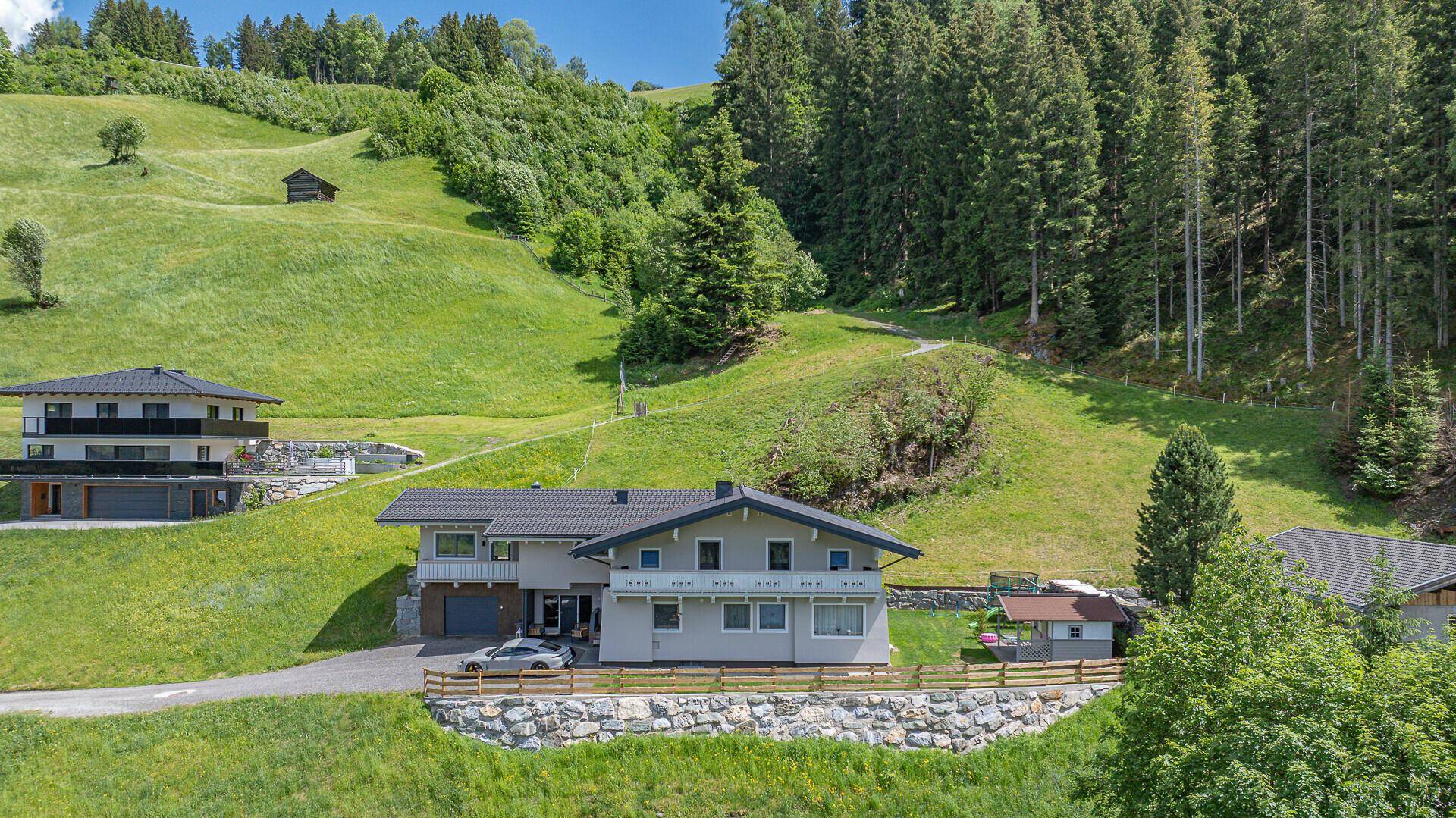 Panoramablick auf zwei Häuser in einer grünen Hügellandschaft mit bewaldeten Bergen im Hintergrund.