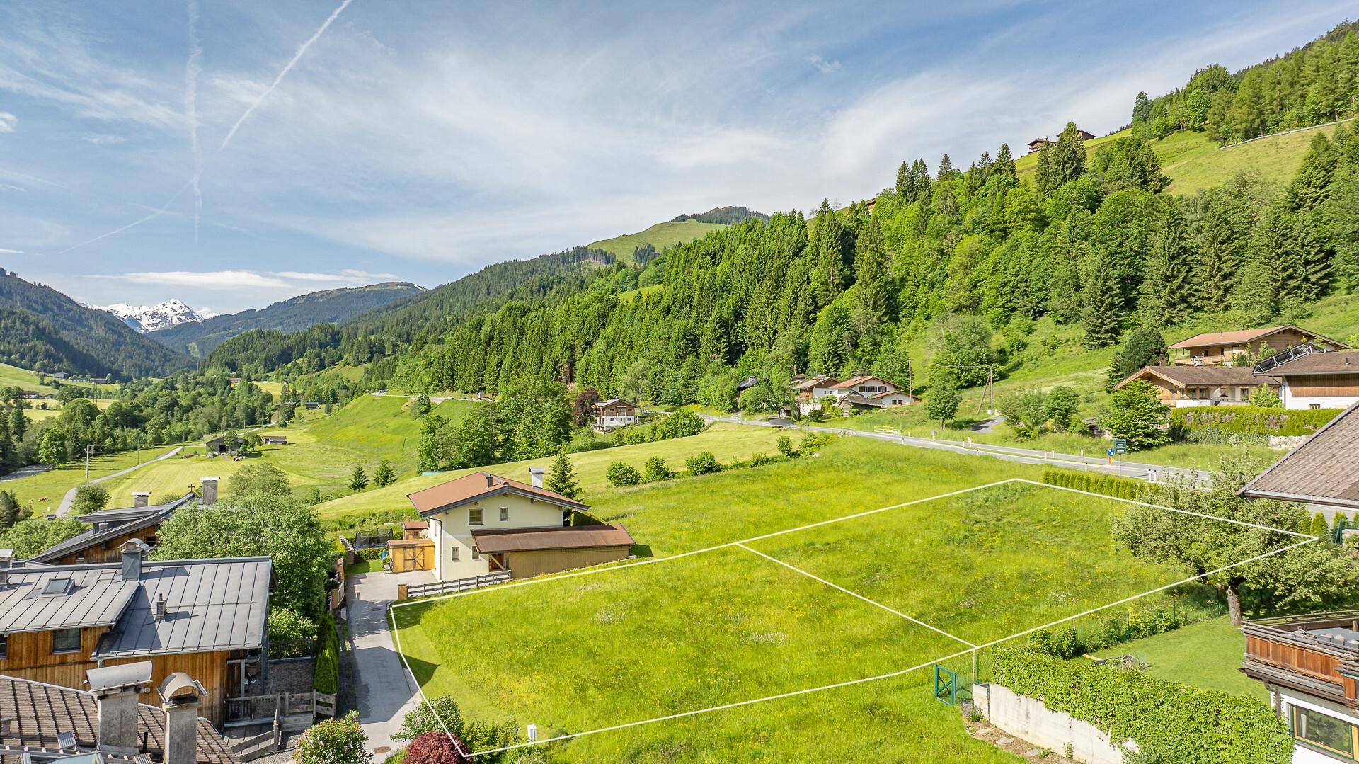 Panoramablick auf ein grünes Tal mit umrandeten Grundstücken und schneebedeckten Berggipfeln im Hintergrund.