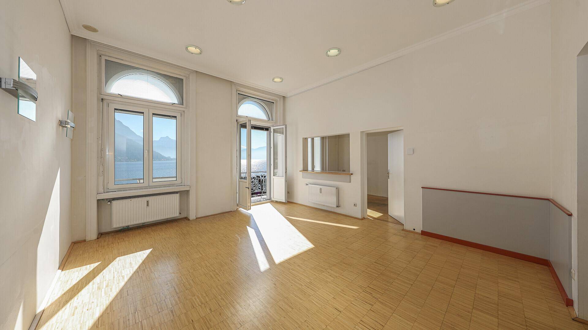 Geräumiger, heller Wohnraum mit Parkettboden und Blick auf den See und die Berge.