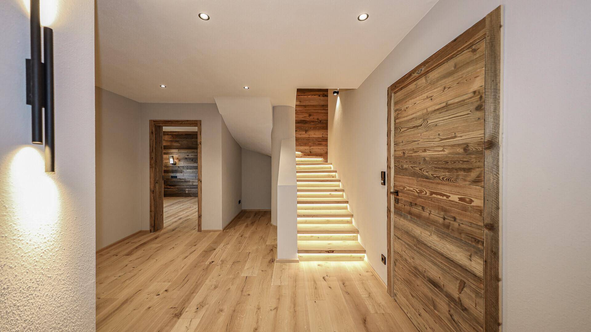 Heller Flur mit beleuchteter Holztreppe und Holzboden, der zu weiteren Räumen führt.