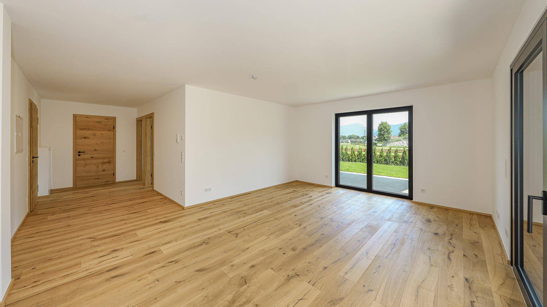 Helles, leeres Zimmer mit Holzboden und Zugang zur Terrasse mit Blick ins Grüne.