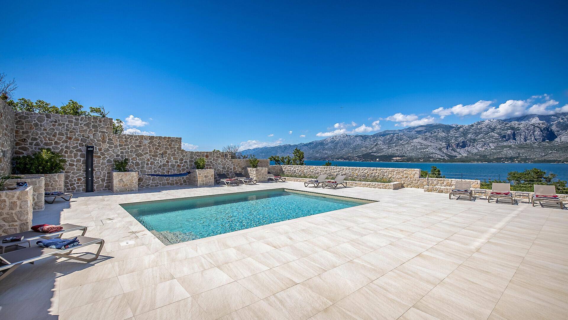 Geräumiger Poolbereich mit hellen Fliesen, bequemen Sonnenliegen und weitem Blick auf das Meer und die Berge.