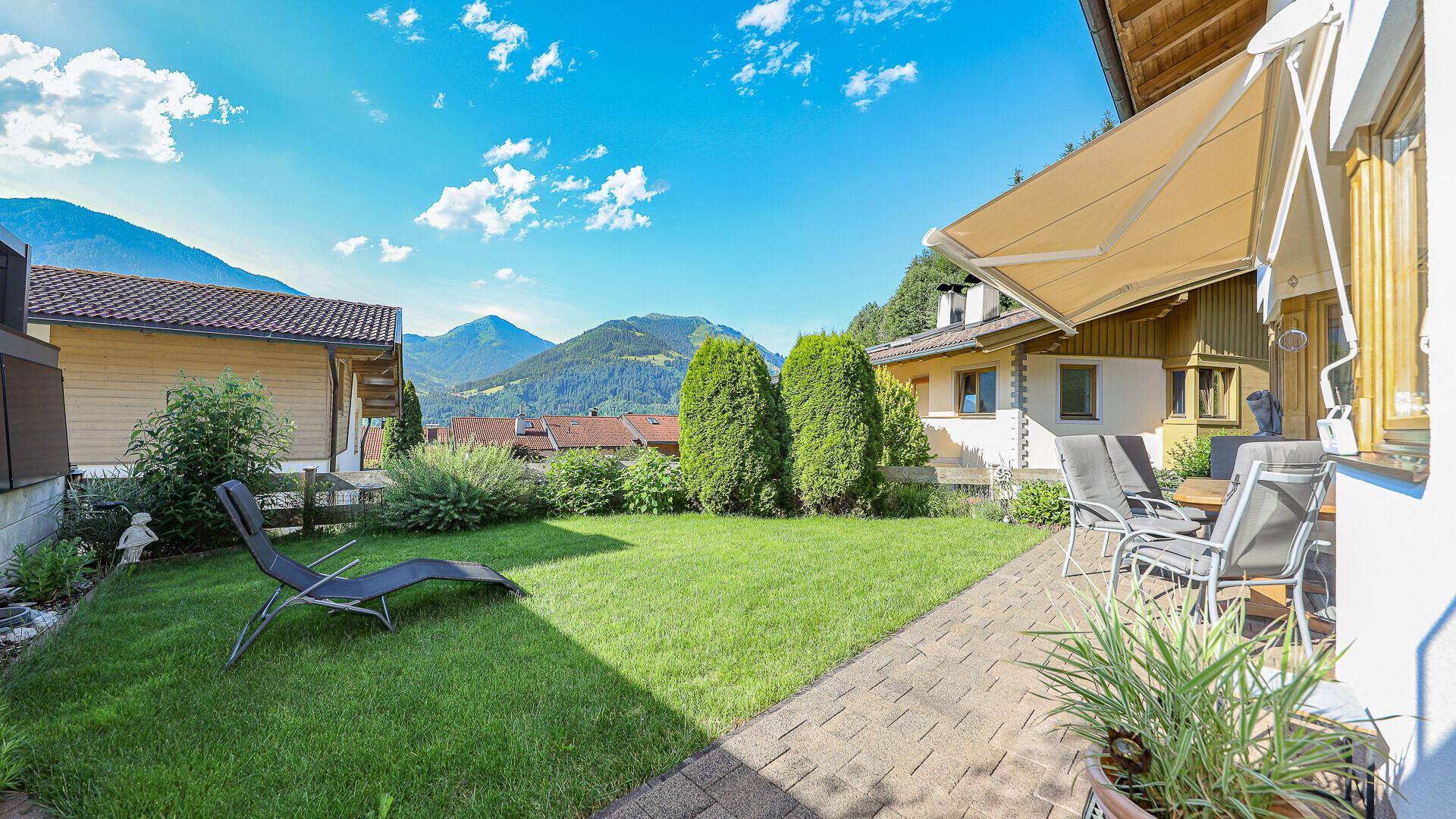 Sonniger Garten mit Rasenfläche, Terrasse und Liegestuhl, umgeben von grüner Natur.