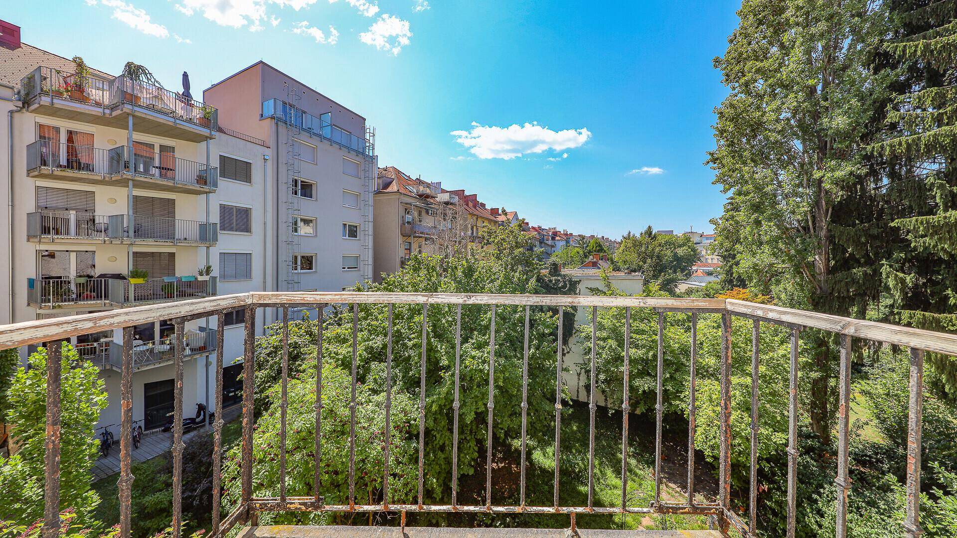 Balkon mit schmiedeeisernem Geländer und Blick auf grüne Bäume und umliegende Gebäude.