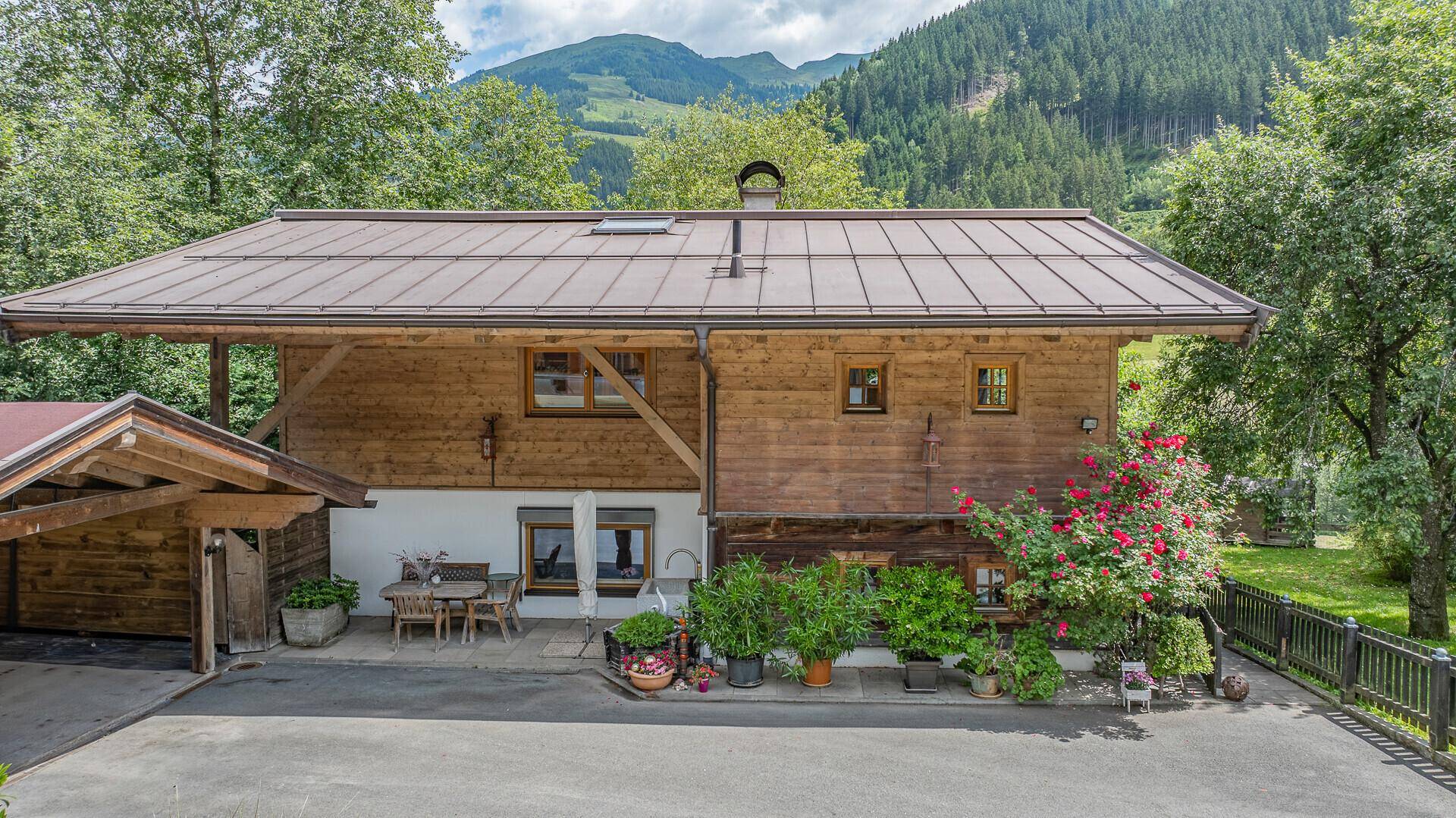 Rückansicht des Holzhauses mit Terrasse, Carport und blühenden Pflanzen im Garten.