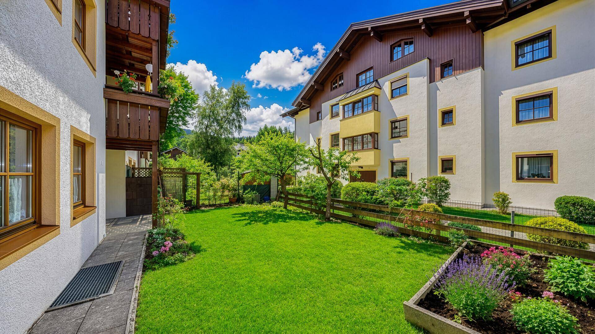 Hausfassade mit Balkonen und gepflegtem Gartenbereich mit Blumenbeeten.