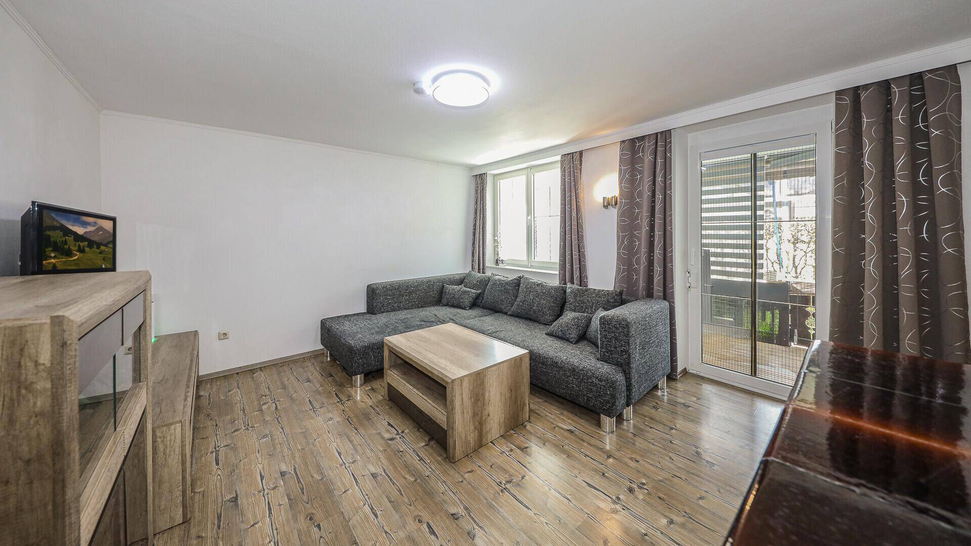 Wohnzimmer mit Holzboden, grauem Sofa und Zugang zu einem Außenbereich durch eine Glastür.