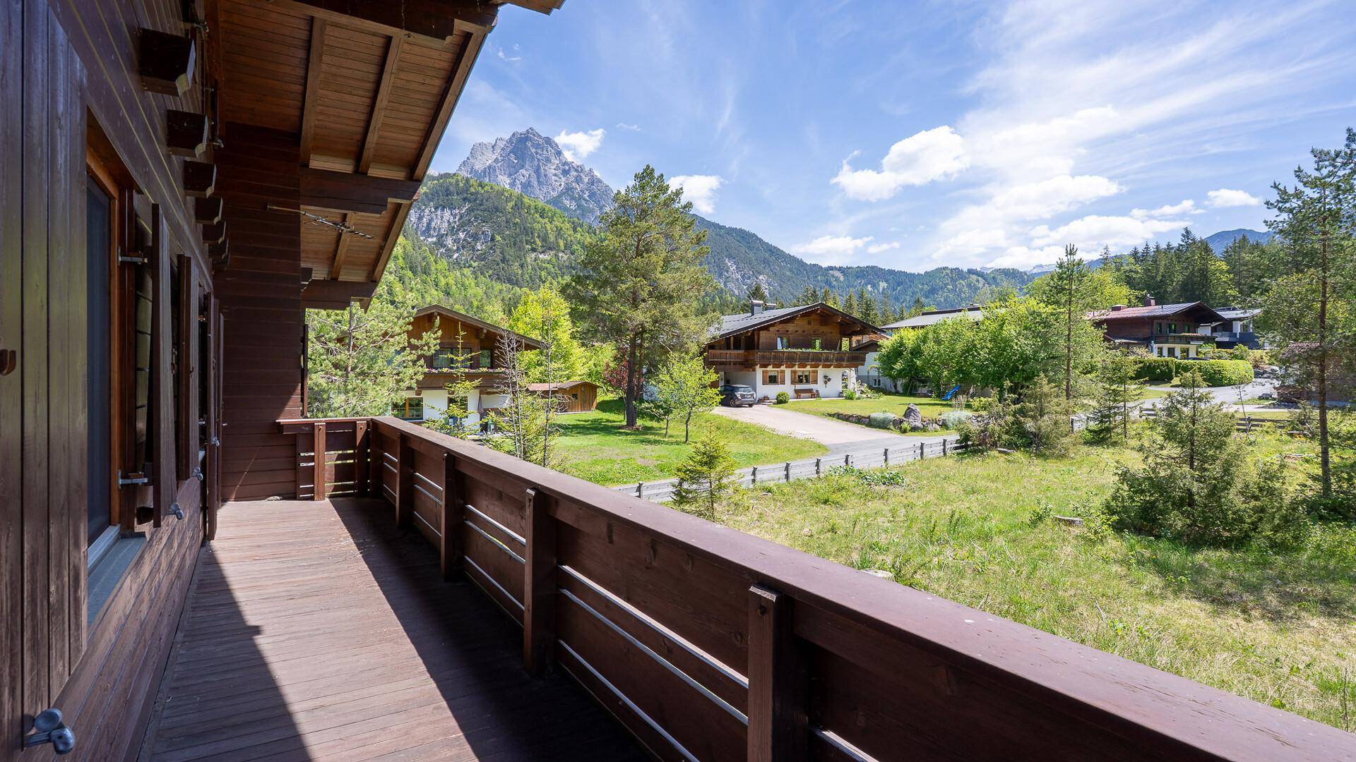 Blick vom Holzbalkon eines Chalets auf grüne Wiesen, Nachbarhäuser und die umliegenden Berge.