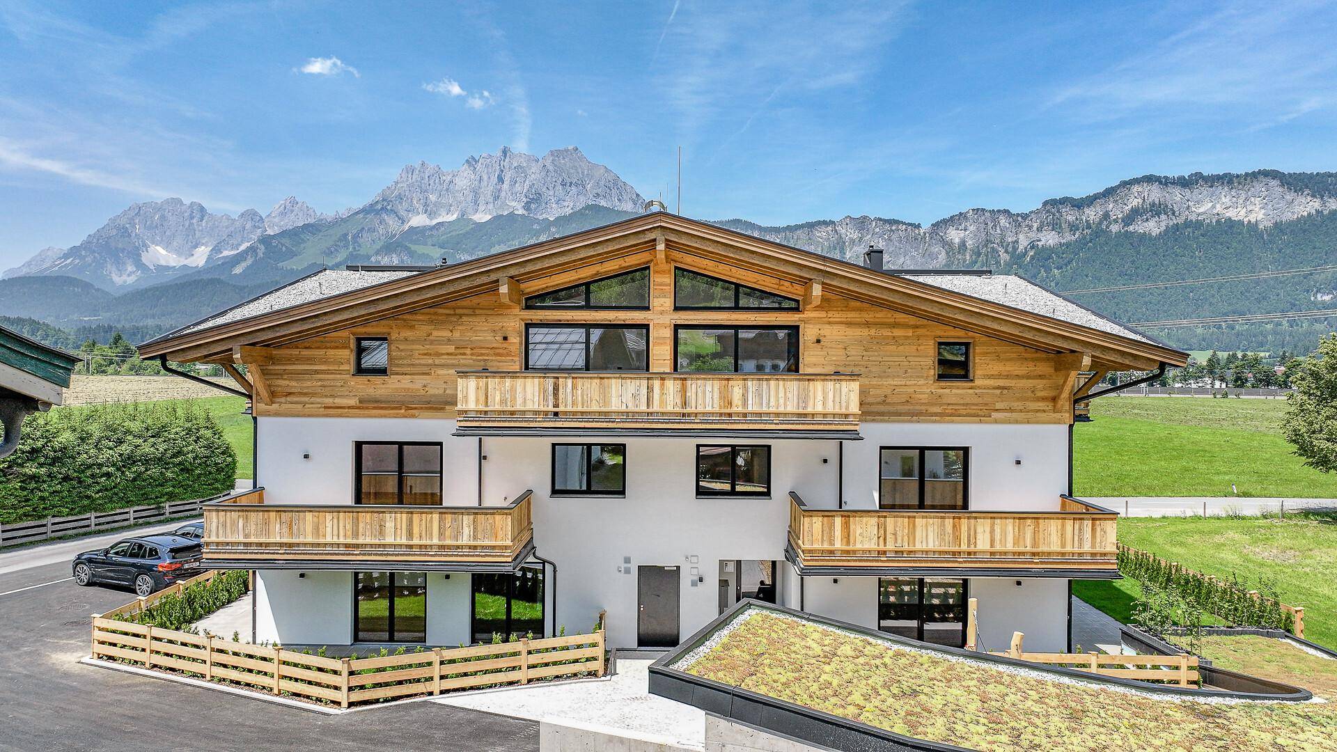 Vorderansicht des modernen Wohnhauses mit Holzelementen und Balkonen vor Bergkulisse.