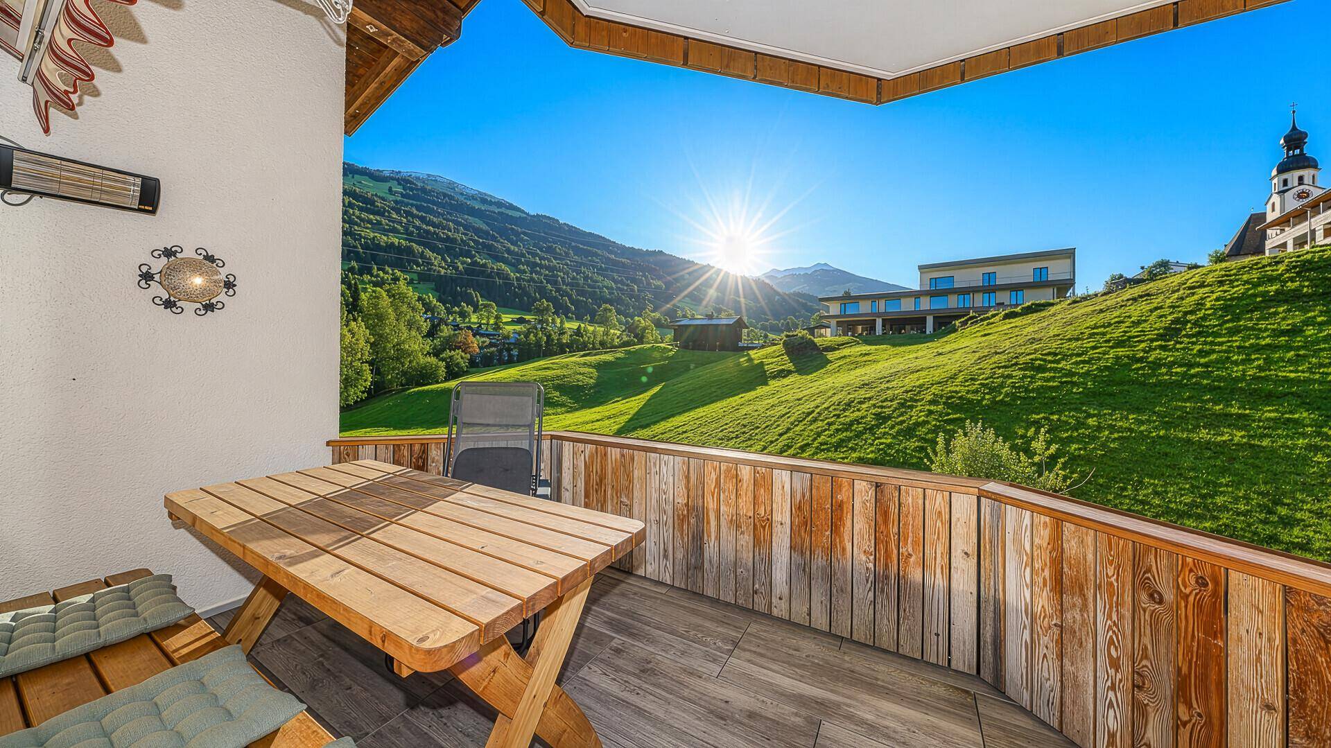 Sonniger Balkon mit Holztisch und Stühlen, bietet einen weiten Blick auf die Berge und grüne Landschaft.