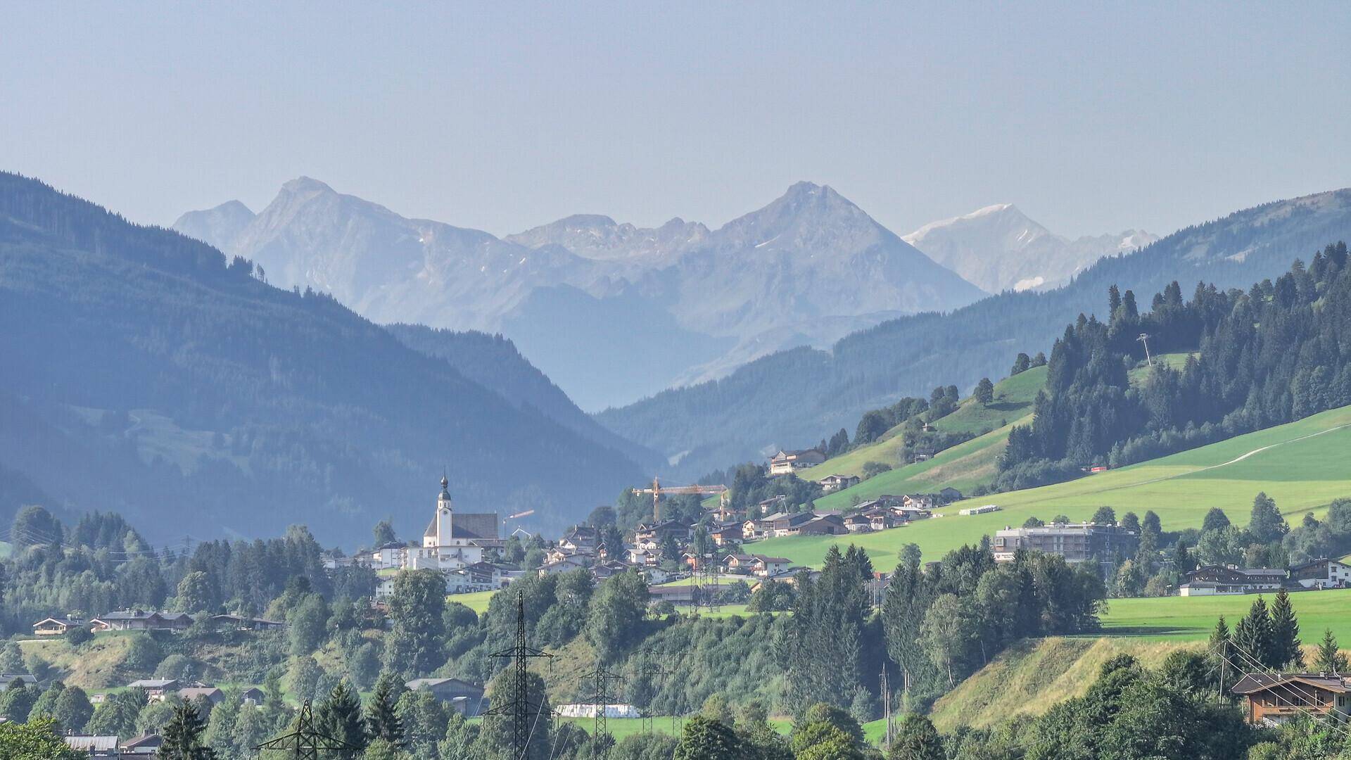 Weitläufiger Blick auf eine malerische Berglandschaft mit einem Dorf und bewaldeten Hügeln im Tal.
