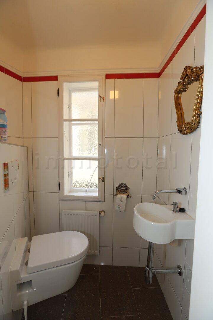 Kleines WC mit Fenster, Wand-WC, Waschbecken und weiß gefliesten Wänden mit rotem Akzent.
