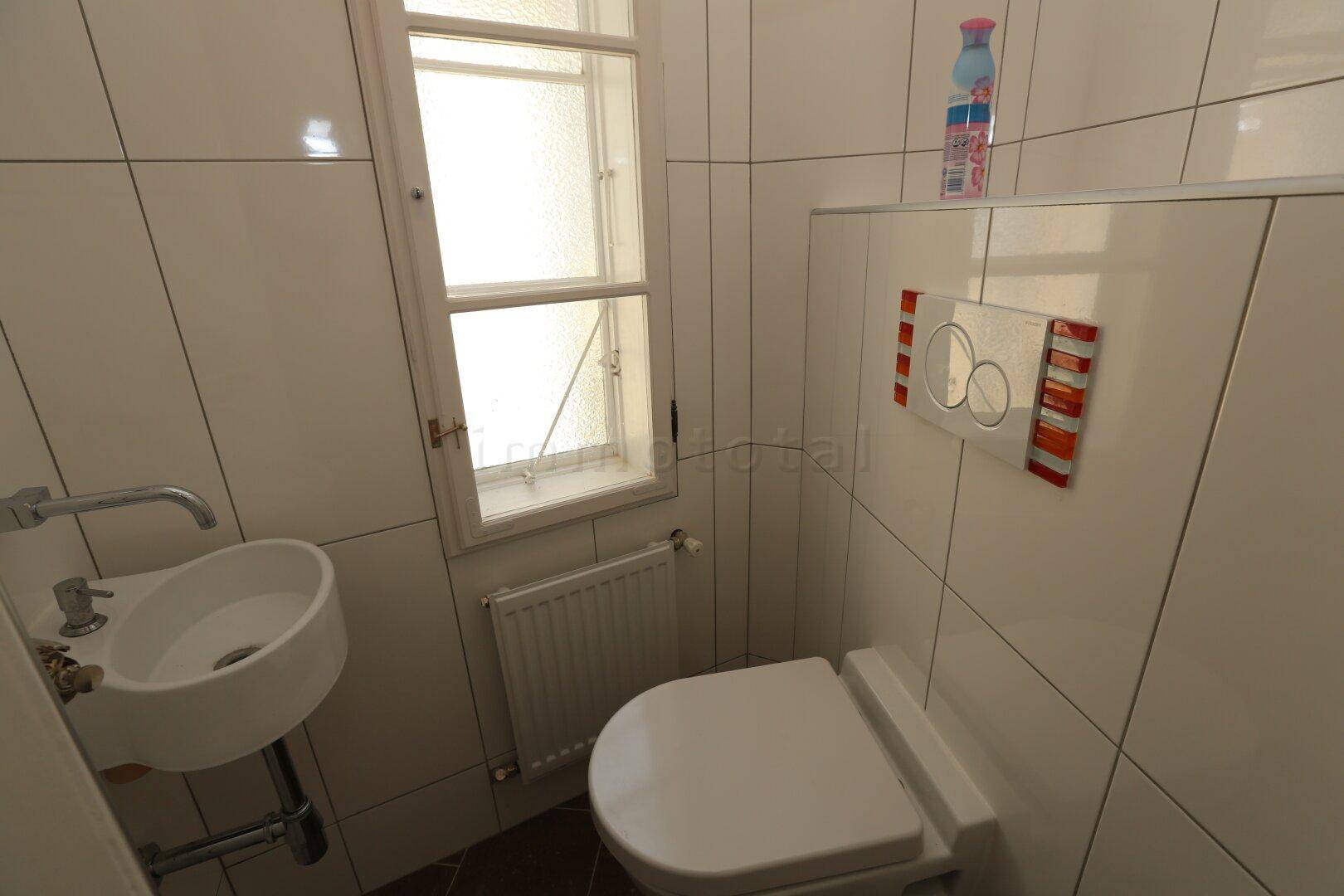 Kleines Gäste-WC mit Fenster, Wand-WC, kleinem Waschbecken und weißen Fliesen.