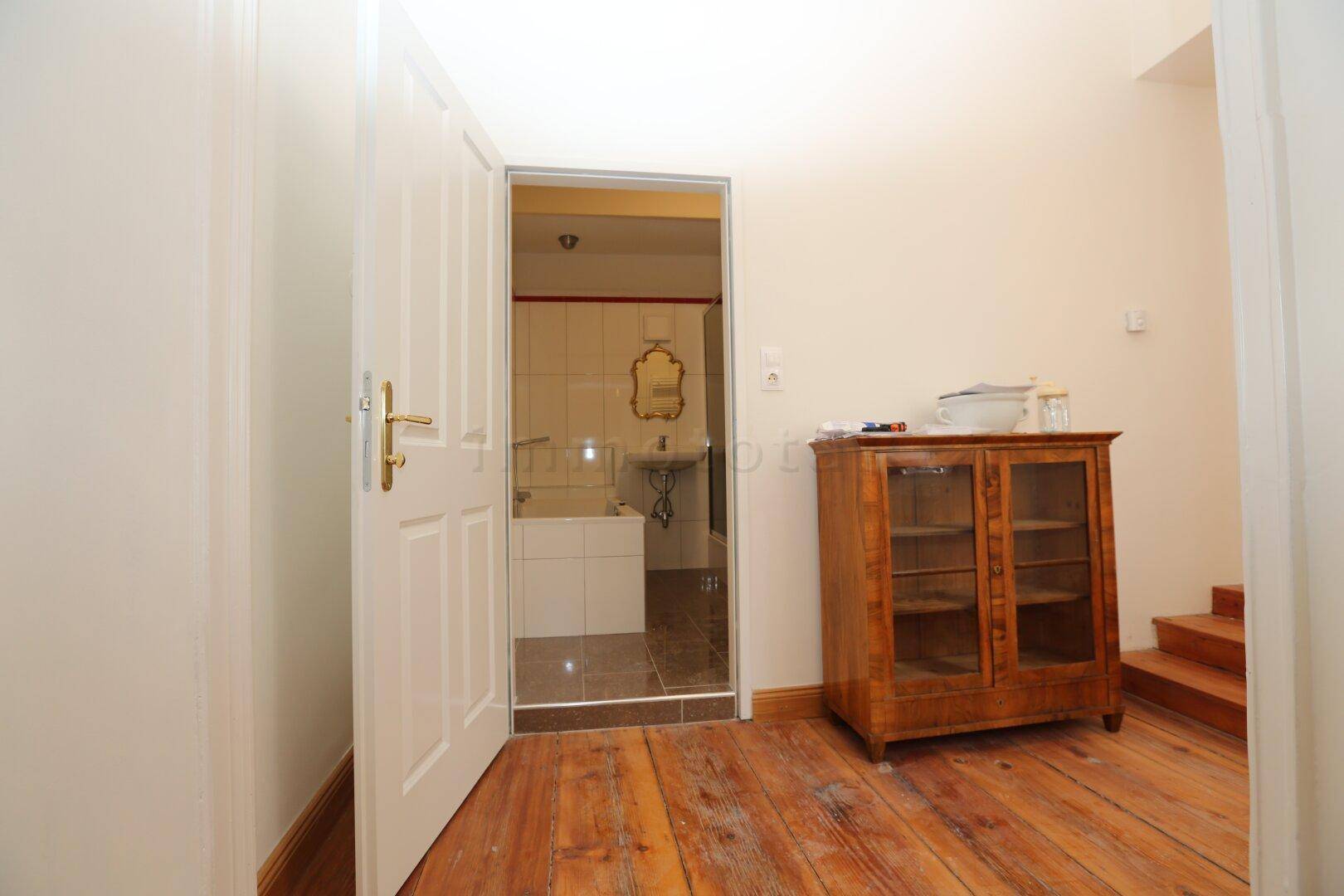 Raum mit Holzdielenboden und antikem Schrank, mit Blick in ein gefliestes WC.