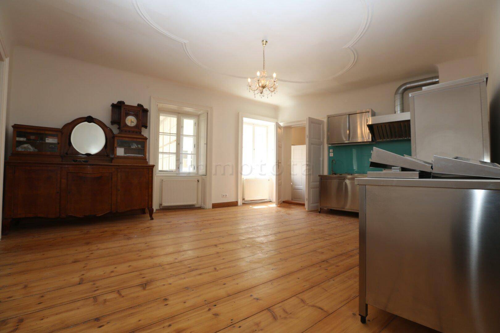 Geräumiger Raum mit Holzdielenboden, Stuckdecke und einem Bereich für eine moderne Edelstahlküche.