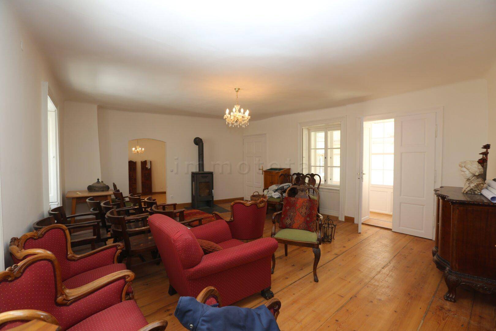 Weitläufiges Wohnzimmer mit Holzdielenboden, Kaminofen und Zugang zu einem weiteren hellen Raum.