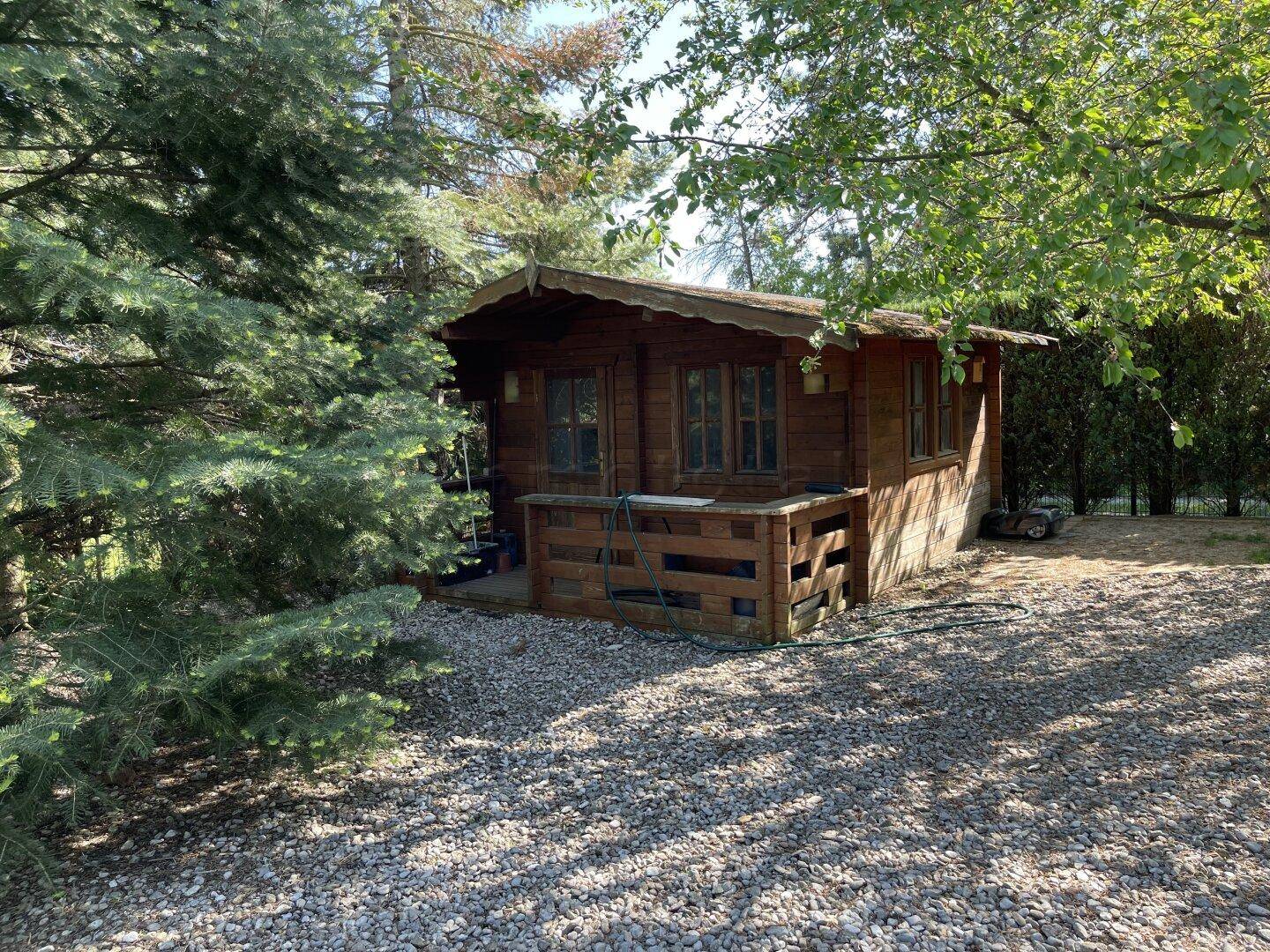 Rustikales Gartenhaus aus Holz, umgeben von Bäumen und einem Kiesweg.