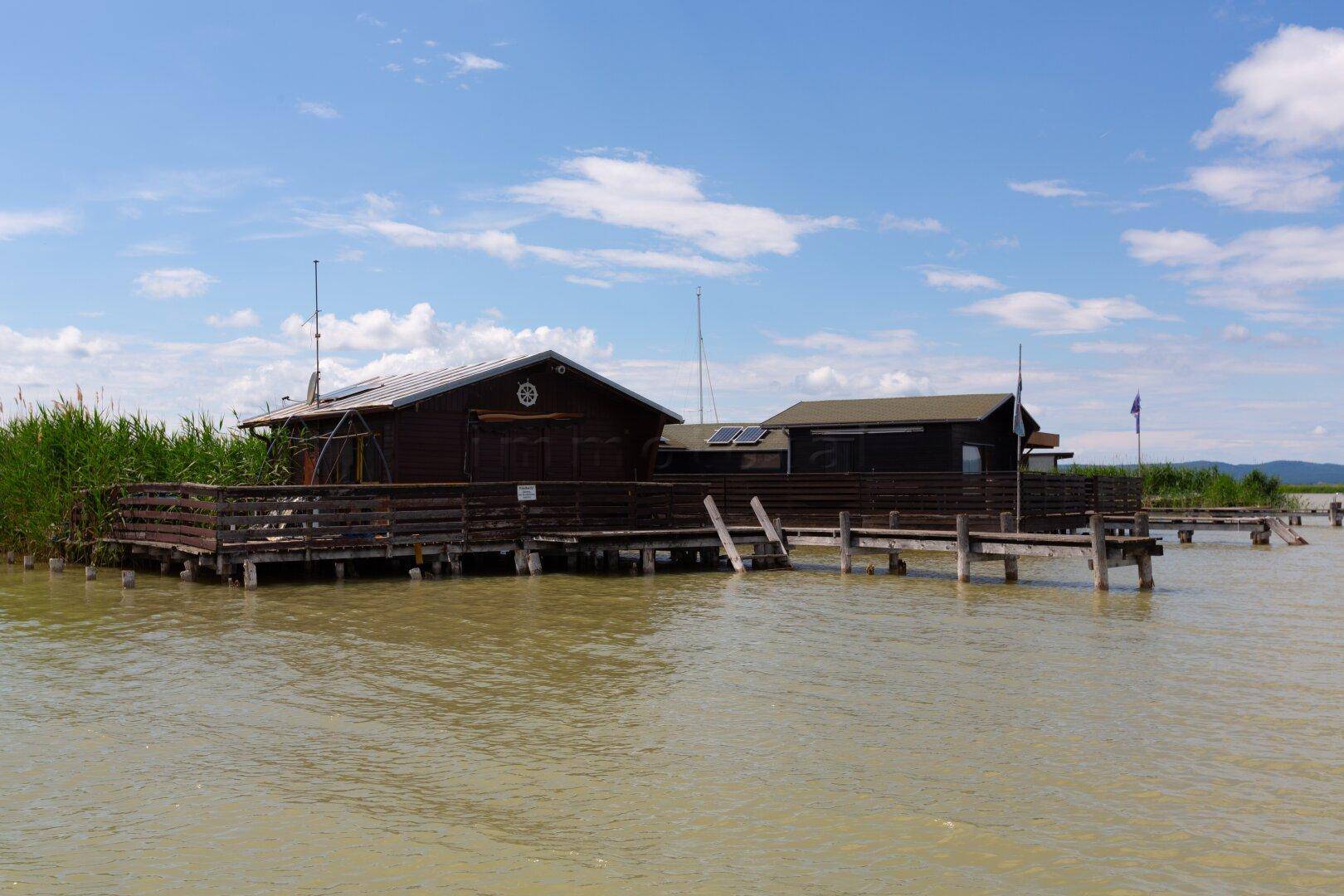 Zwei rustikale Holzhäuser auf Stelzen über dem Wasser, verbunden durch eine Holzterrasse mit Schilf.