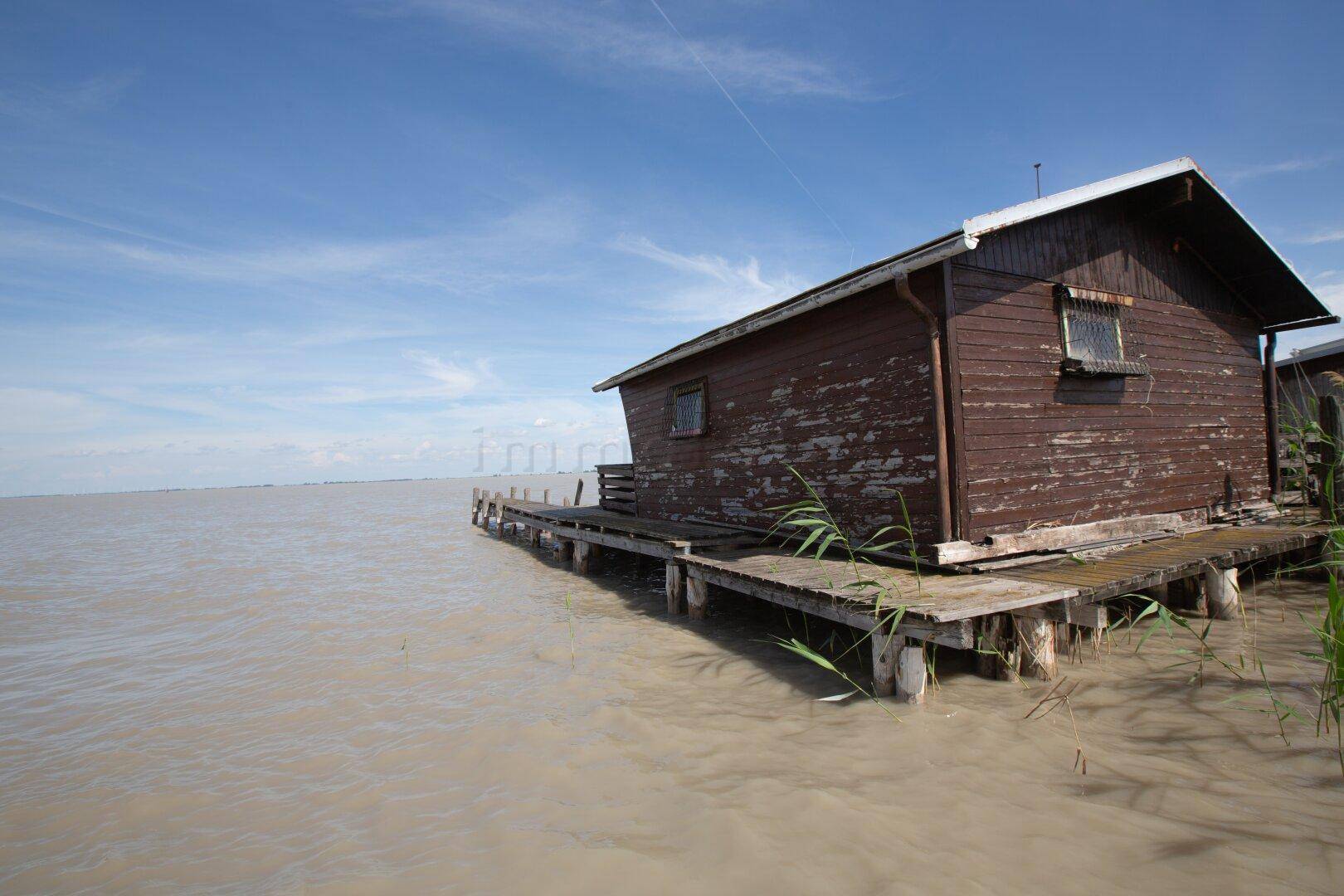 Seitenansicht eines Holzhauses auf Stelzen über dem Wasser mit einem langen Holzsteg in den See.