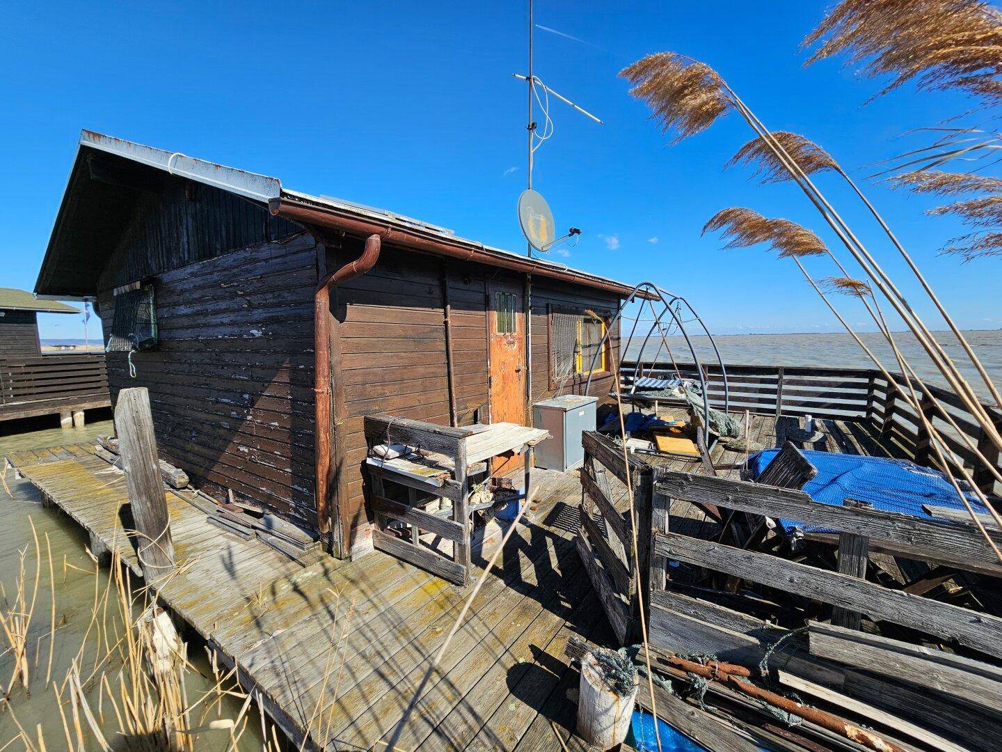 Hölzerne Terrasse mit Outdoor-Möbeln, einem kleinen Kühlschrank und Blick auf das Wasser und Schilf.