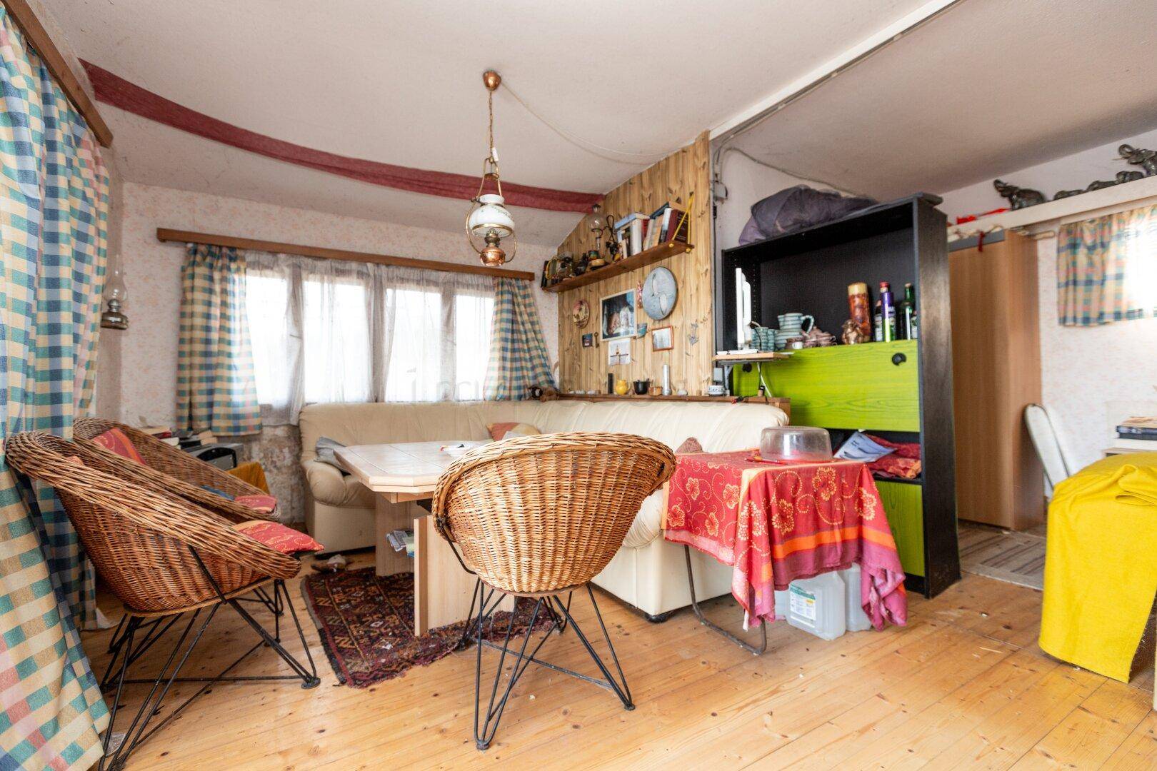 Gemütlicher Wohn-Essbereich mit Holzboden, Korbstühlen, Sofa und einem Esstisch unter einem Fenster.