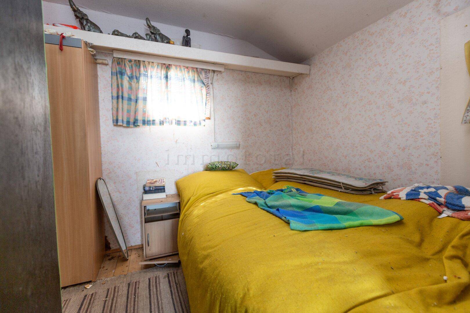 Einfaches Schlafzimmer mit gelbem Bett, kleinem Fenster und einem Kleiderschrank in rustikalem Stil.