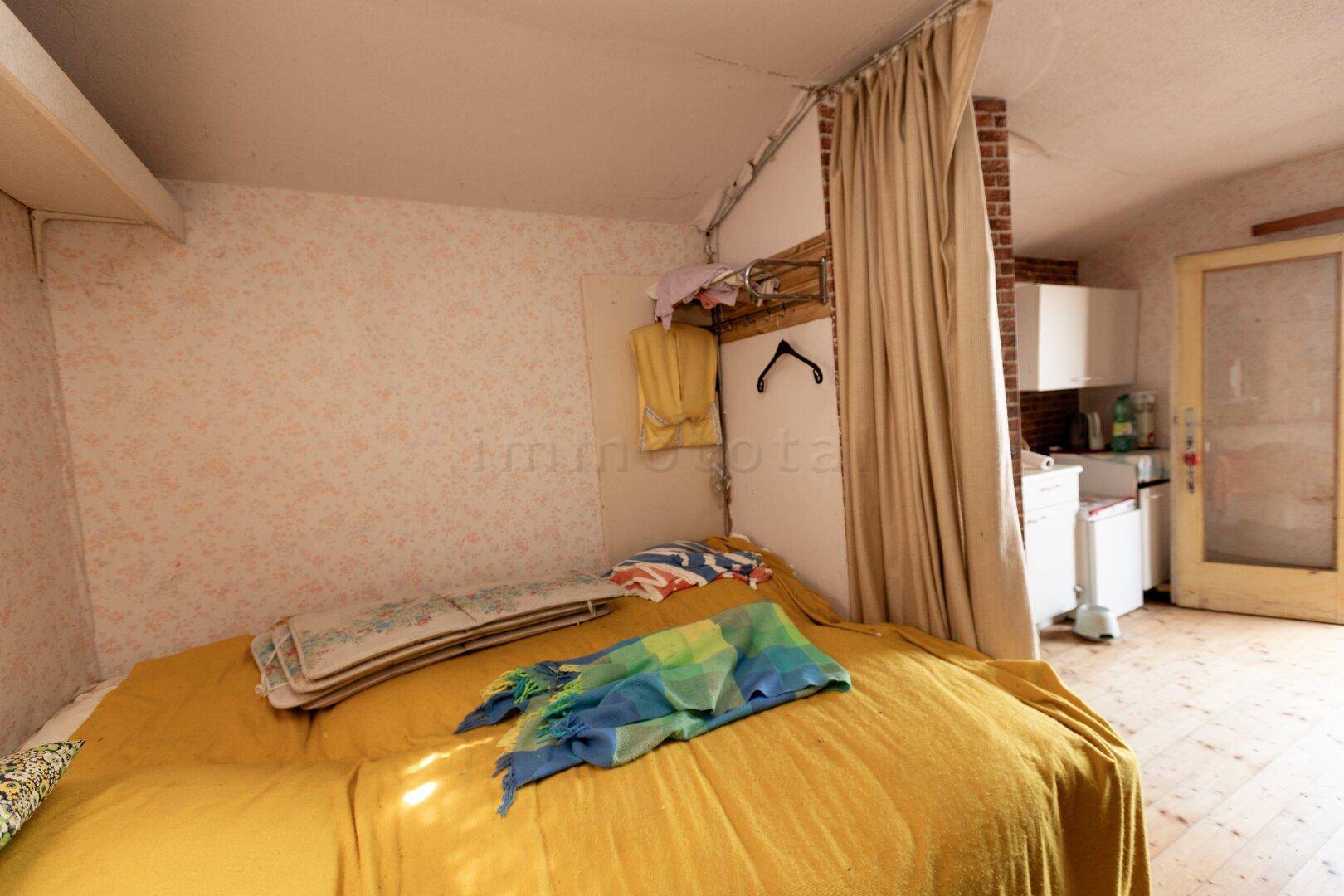 Schlafzimmer mit gelbem Bett, durch einen Vorhang vom Küchenbereich getrennt, mit Holzboden.
